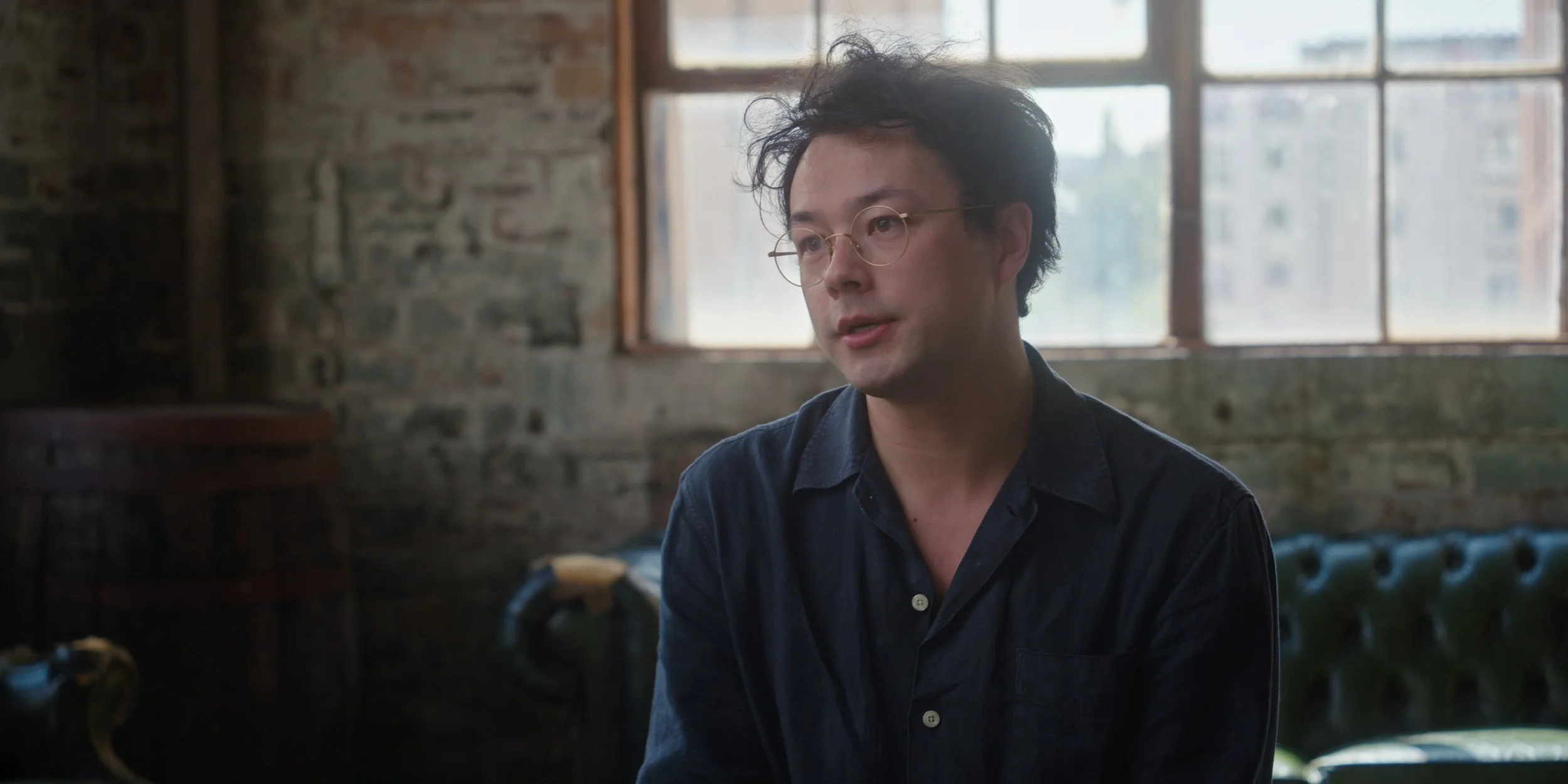 A man with curly black hair, glasses, and a dark shirt sitting in a rustic room with large windows and an exposed brick wall.