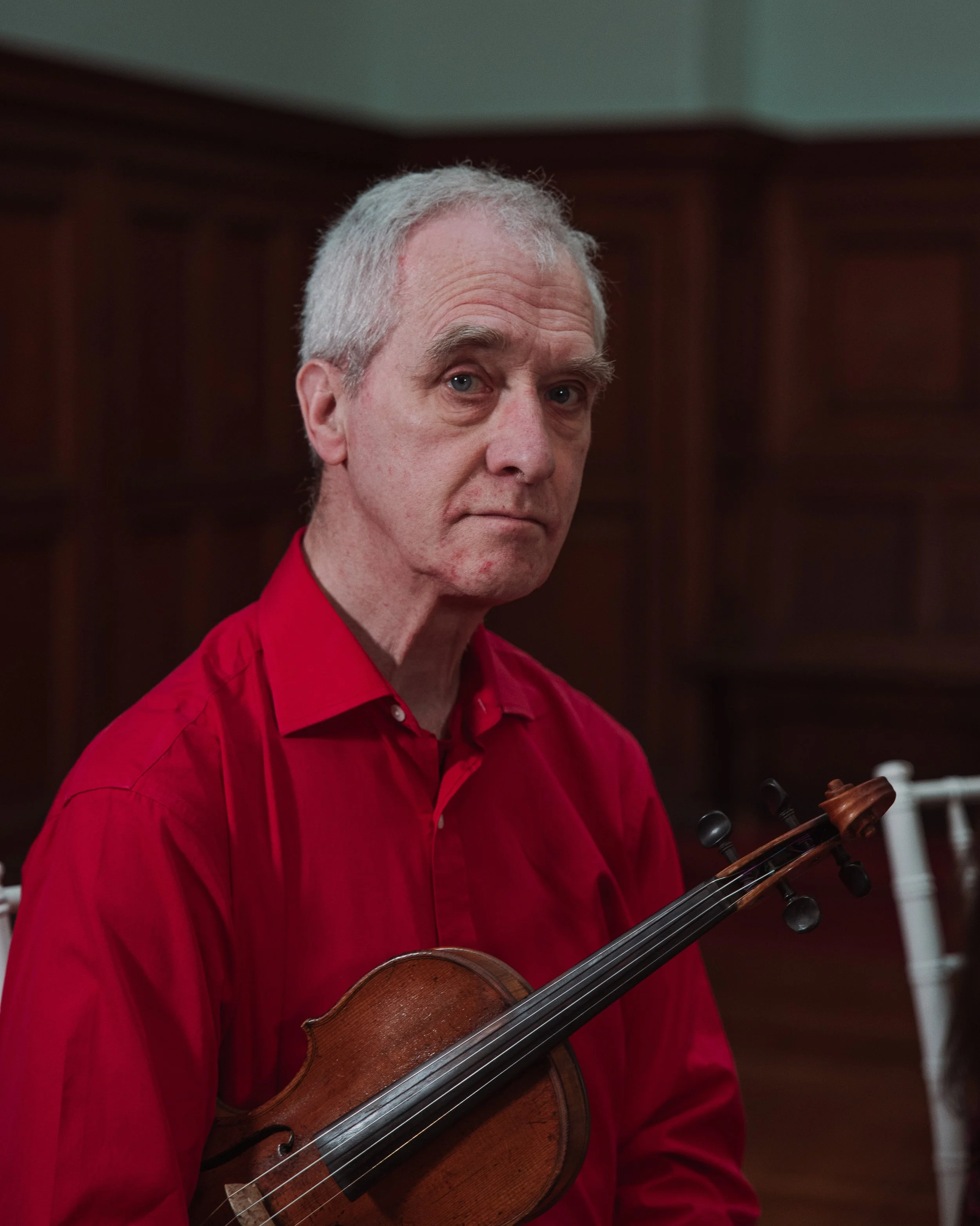 An elderly man with gray hair and a serious expression, wearing a red shirt, holding a violin, seated indoors with dark wood paneling in the background.