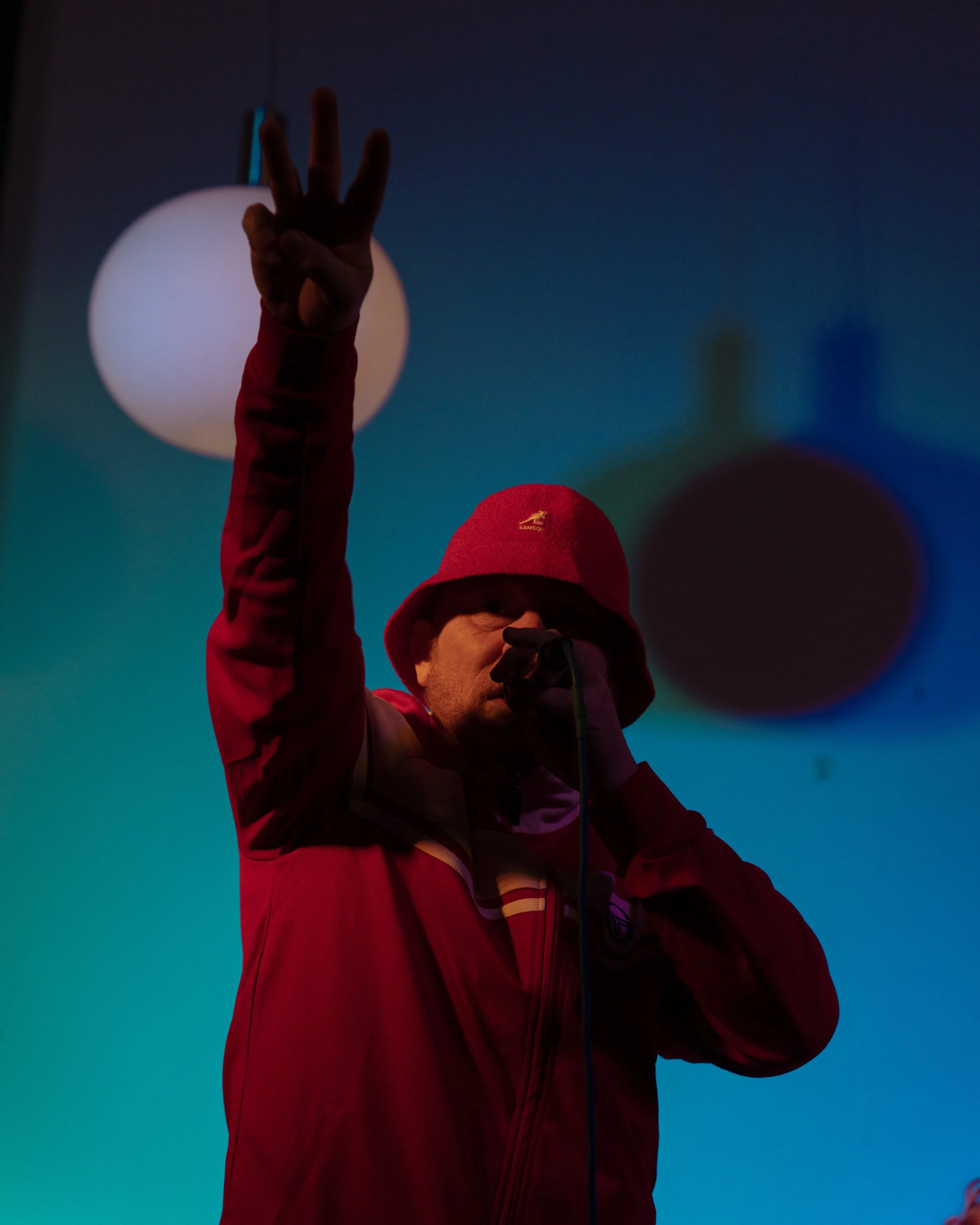 Person wearing red jacket and red hat, holding microphone, making a peace sign with their right hand, indoors with blue and white spherical lights in the background.