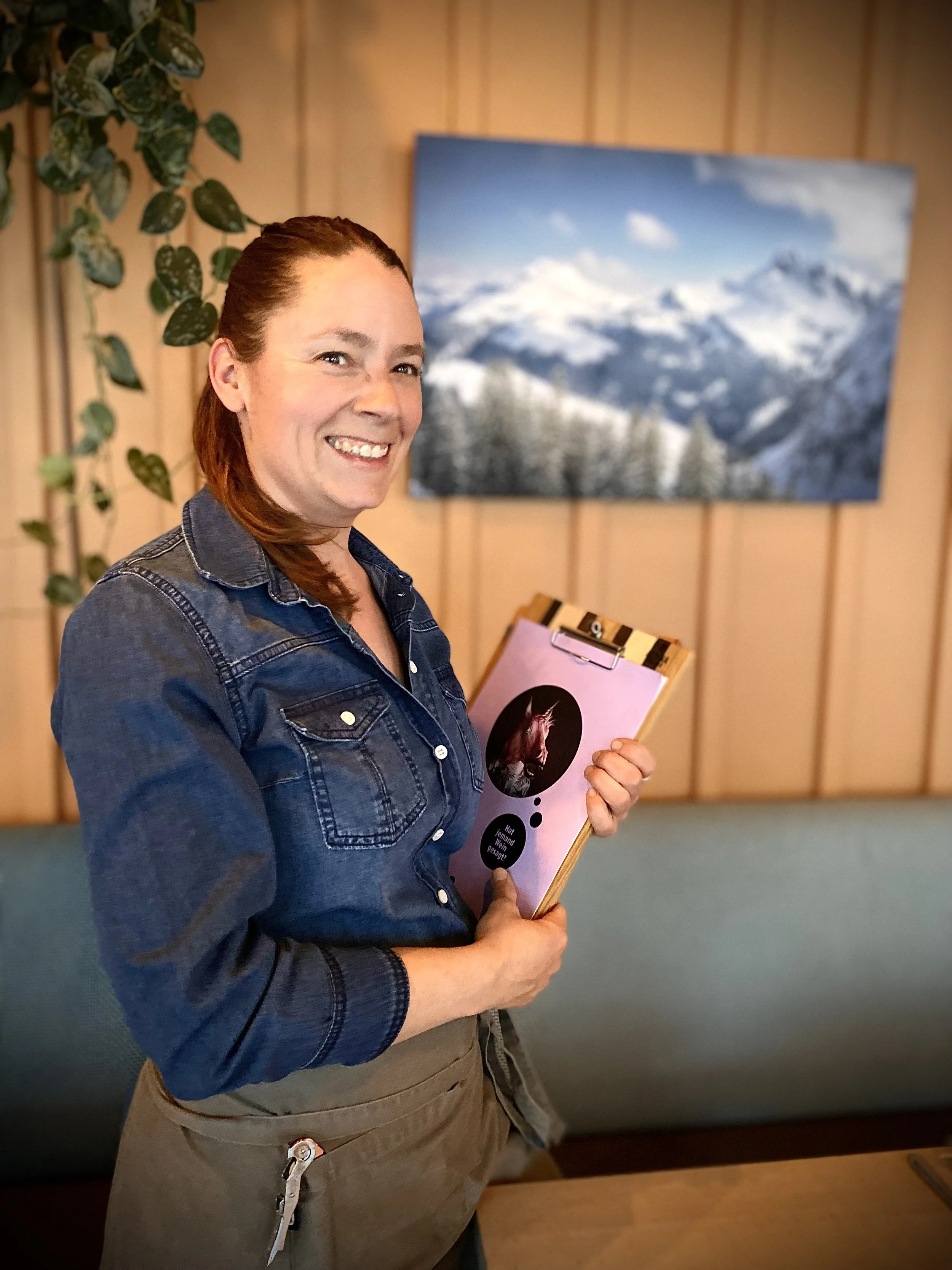 Frau in Jeanshemd mit Klemmbrett, steht vor einem Wandbild mit Berglandschaft und Pflanzen im Hintergrund.