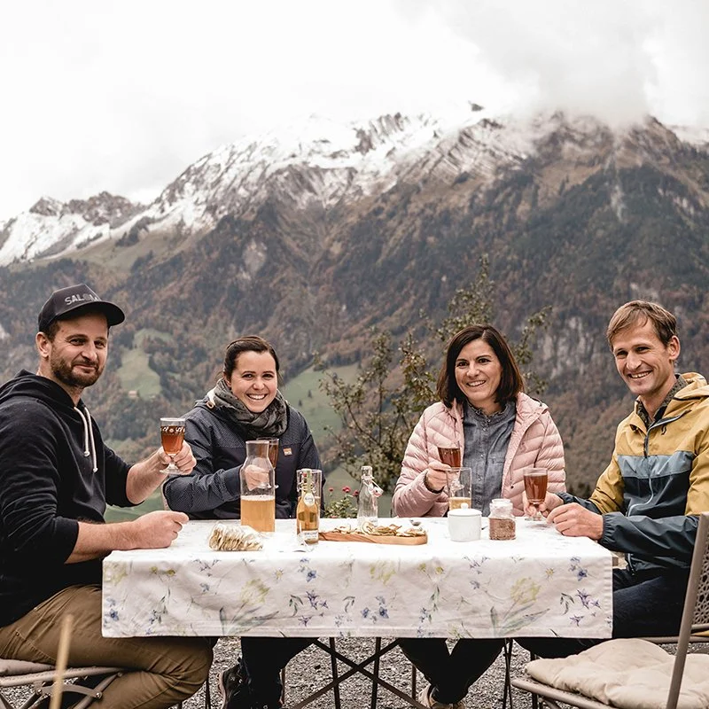 Vier Personen sitzen an einem Tisch im Freien und trinken Getränke. Im Hintergrund sind verschneite Berge zu sehen. Sie lächeln und genießen die Landschaft.