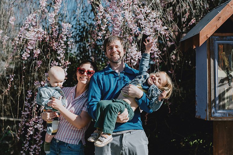 A family of four wearing pink and blues smiles from beneath a canopy of cherry blossoms during a family photo session in Asheville, NC