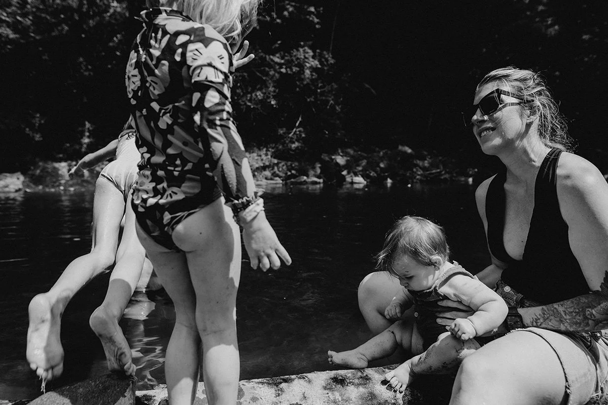 a family smiles on a riverbank. The photo is in black and white and made by the top documentary family photographer in the world, Melina Coogan