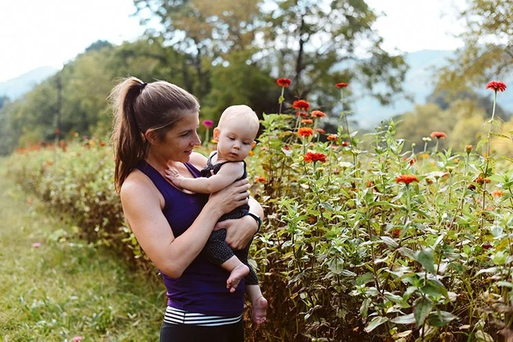A mom with a pony tale holds a baby girl in a field of flowers during a family photo shoot in Asheville North Carolina