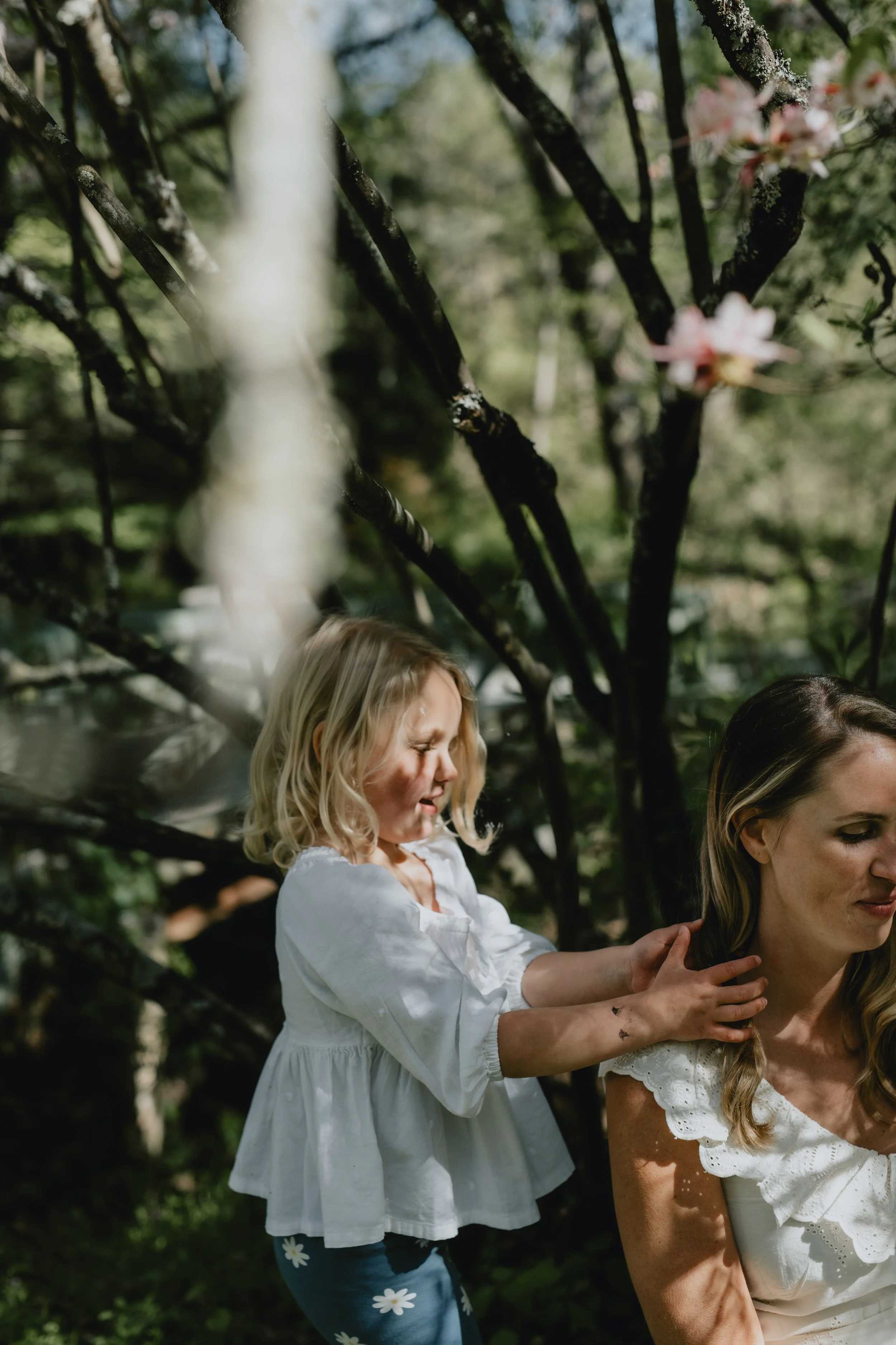 A four year old girl plays with her mother's hair with dappled light. There are pale, spring like flowers and the girls are wearing white dresses during a family photo shoot in Asheville.