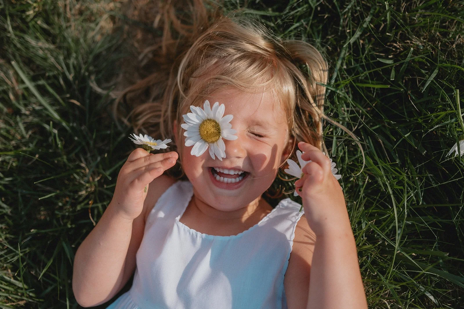 A young girl with blonde hair lying in grass, smiling with her eyes closed, holding white daisies near her face.