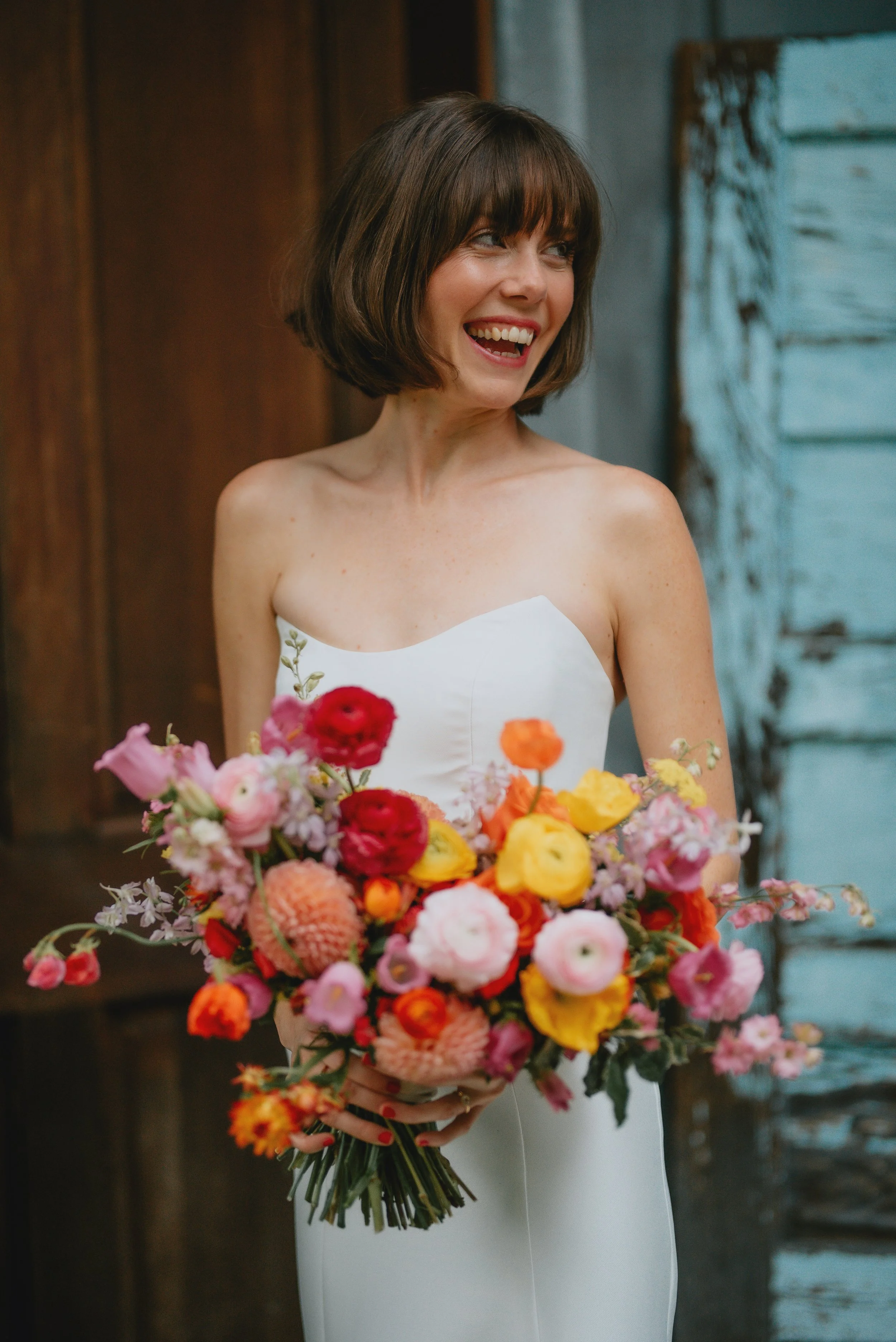 A woman with a short brown bob haircut, smiling, wearing a strapless white dress, holding a large colorful bouquet of flowers in front of her with a rustic wooden background.