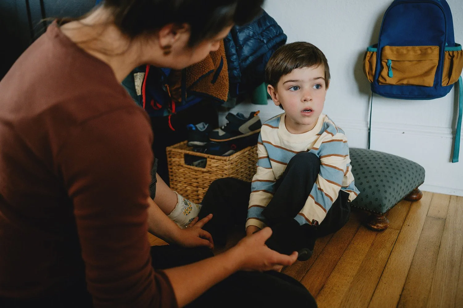 a little boy sits on the floor of his home as his mom helps him put his shoes on.