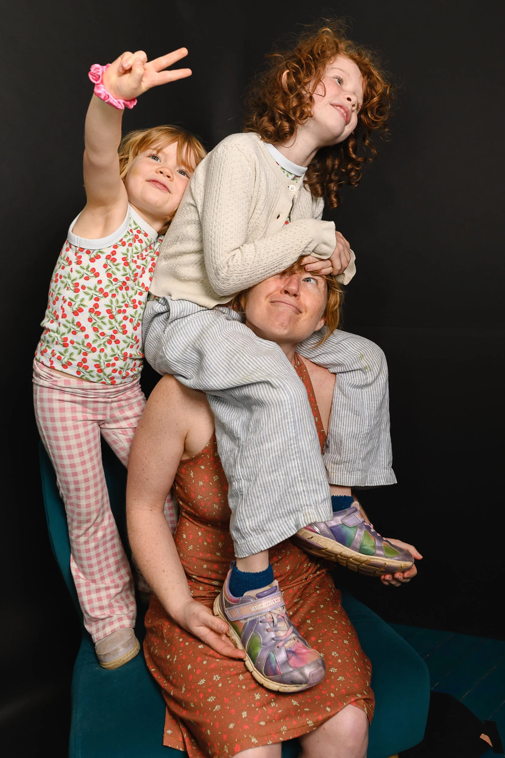 A woman sitting on a chair with two children on her shoulders, one girl with curly red hair and the other girl with straight red hair, making peace signs and smiling against a black background.
