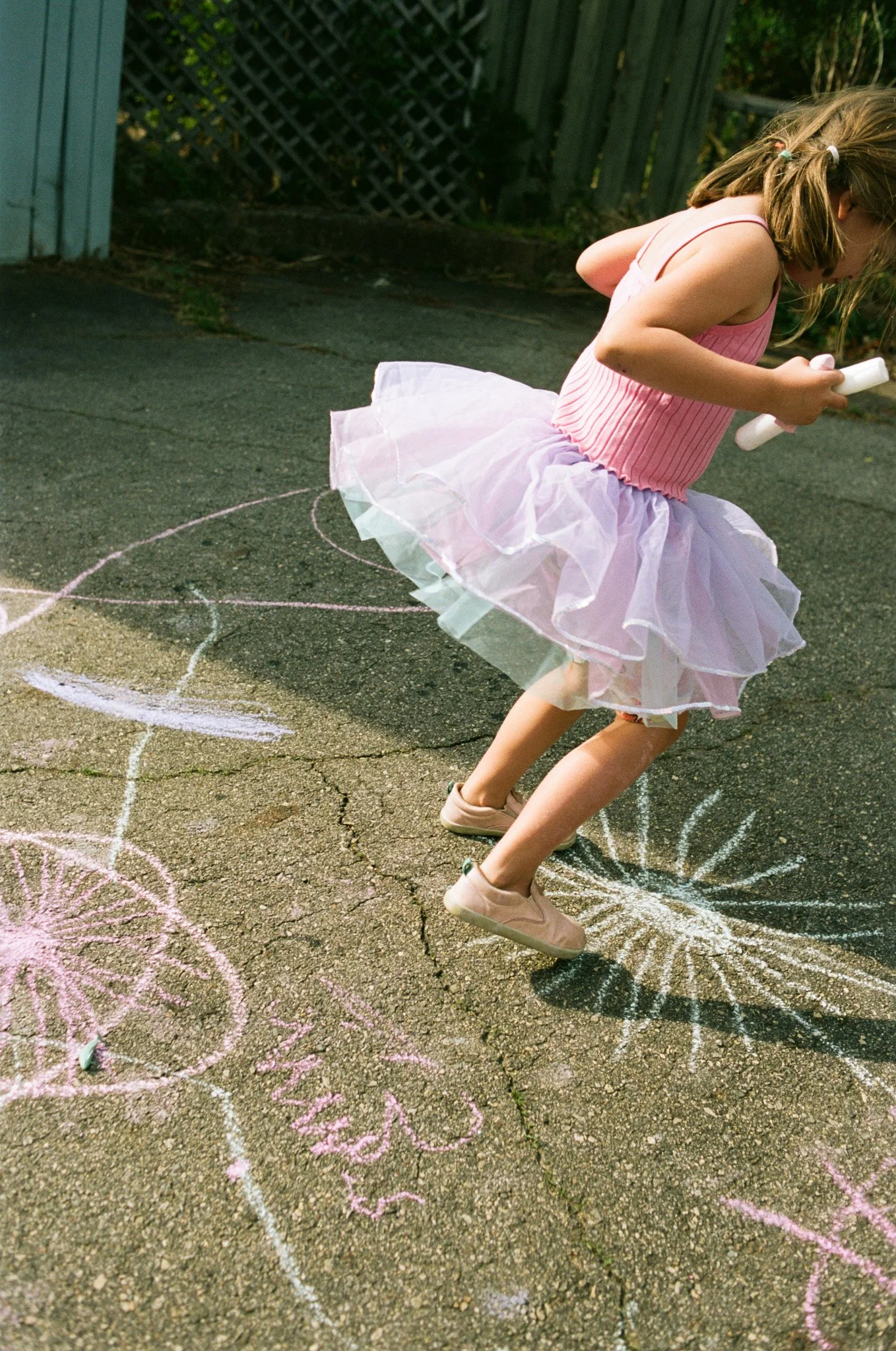A girl with a purple skirt and pink tank top jumps out of the frame. She is holding chalk and jumping through a driveway filled with chalk art during a family photo session.