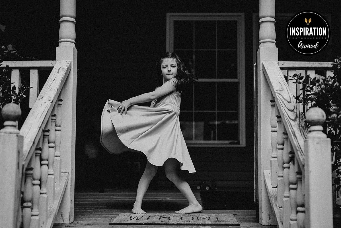 A young girl in a dress dancing or posing on a porch with wooden stairs and railings, front door behind her, in black and white.