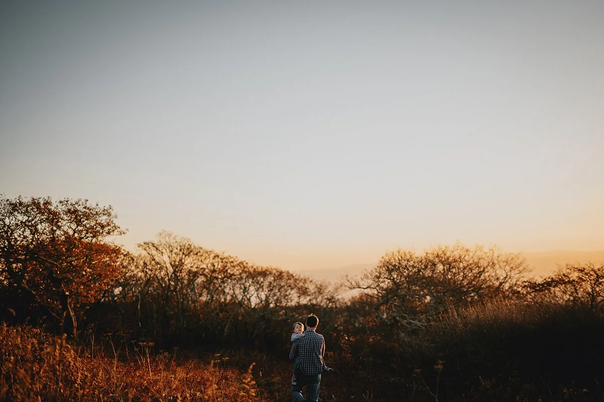 A sweeping mountaintop view at sunset, a man carries his little boy during a family photo shoot in Asheville, NC