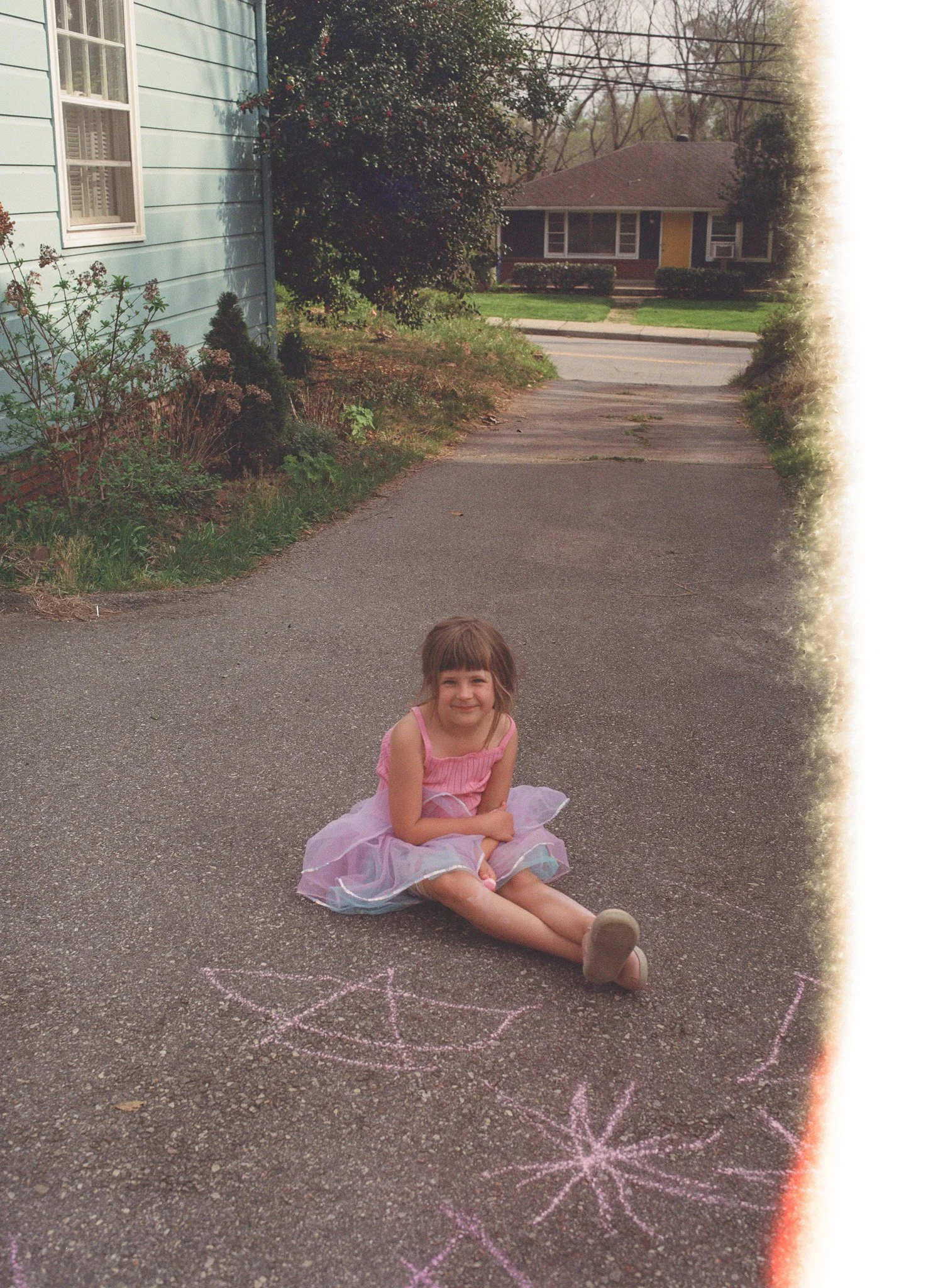a six year old girl in a purple fairy skirt smiles at the camera inside her driveway on a film image, there is al ight leak on the right side of the image.