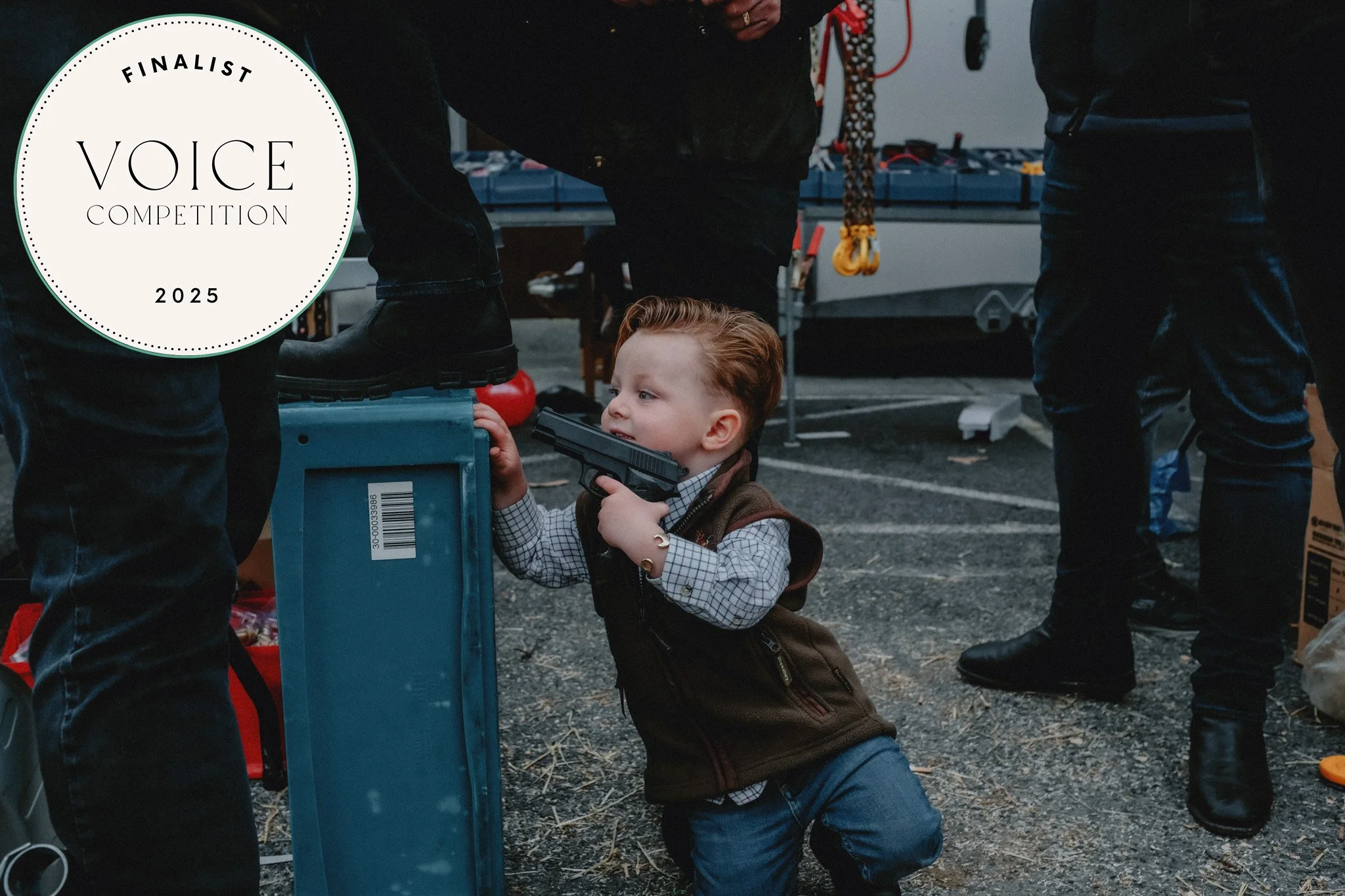 A young boy with red hair, wearing a checkered shirt and brown vest, kneels on the ground holding a toy gun to his mouth in an outdoor setting with people around, some standing and some on a work truck, at a contest or event.