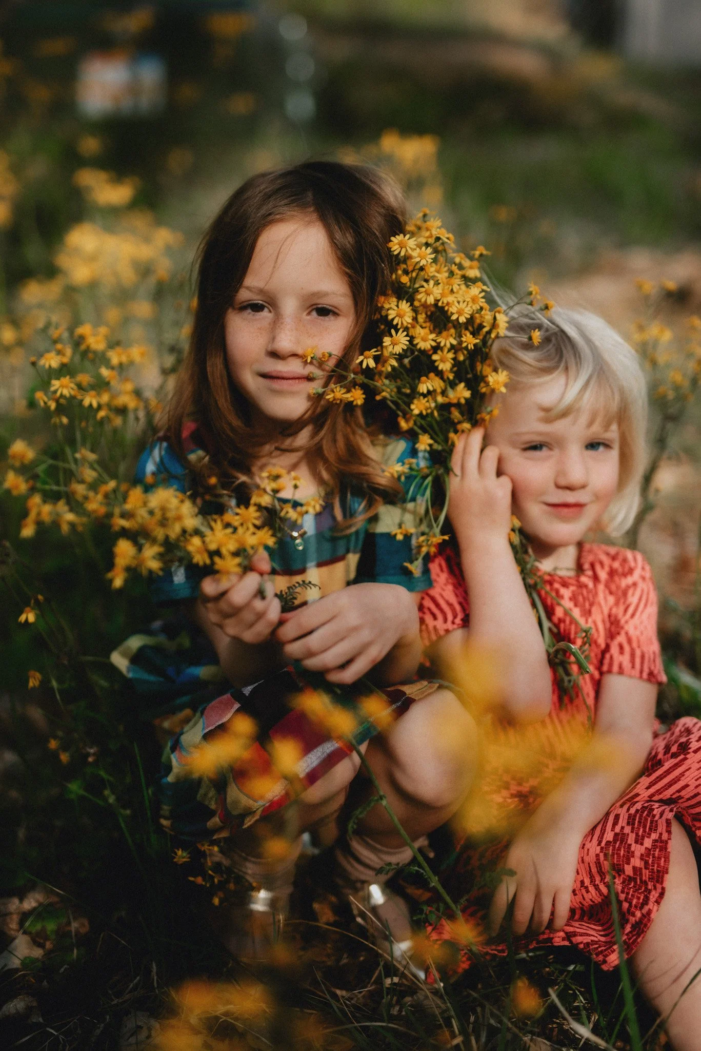 two young girls pose for a photo amidst a field of bright yellow flowers. They are wearing bold, vivid colors and holding bunches of yellow daisies.