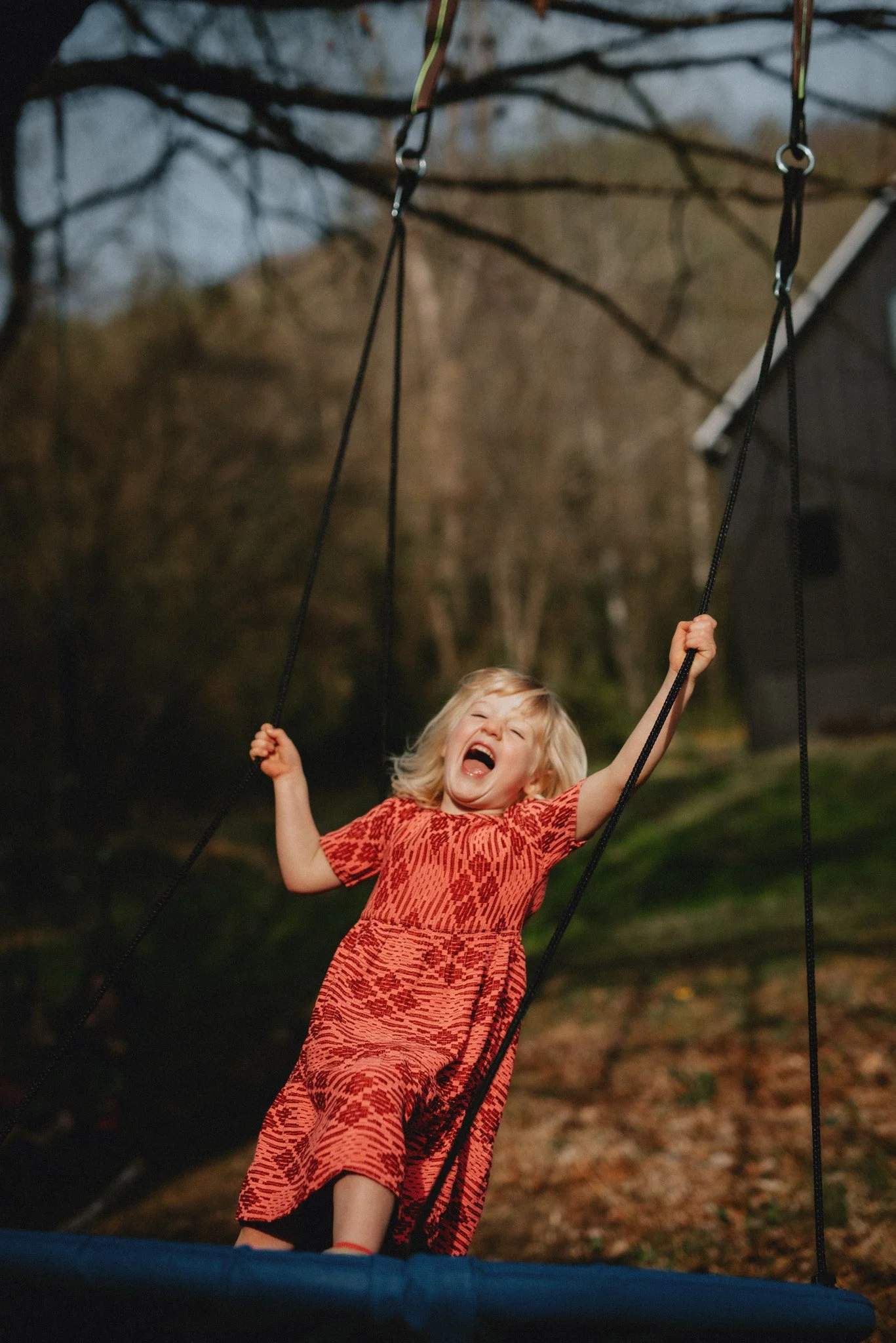 A four year old girl in a bright red dress yells in excitement and joy while on a backyard swing during a spring evening.