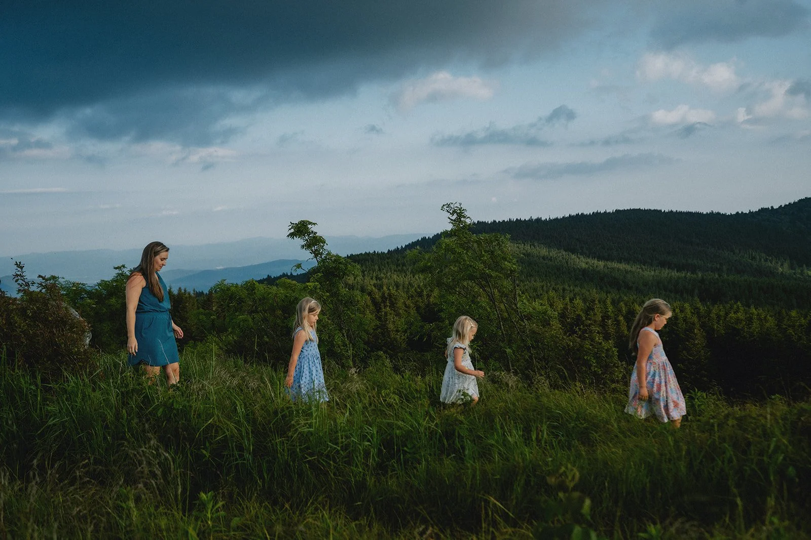 A woman and four young girls walking in a grassy field with rolling hills and dark clouds in the sky.