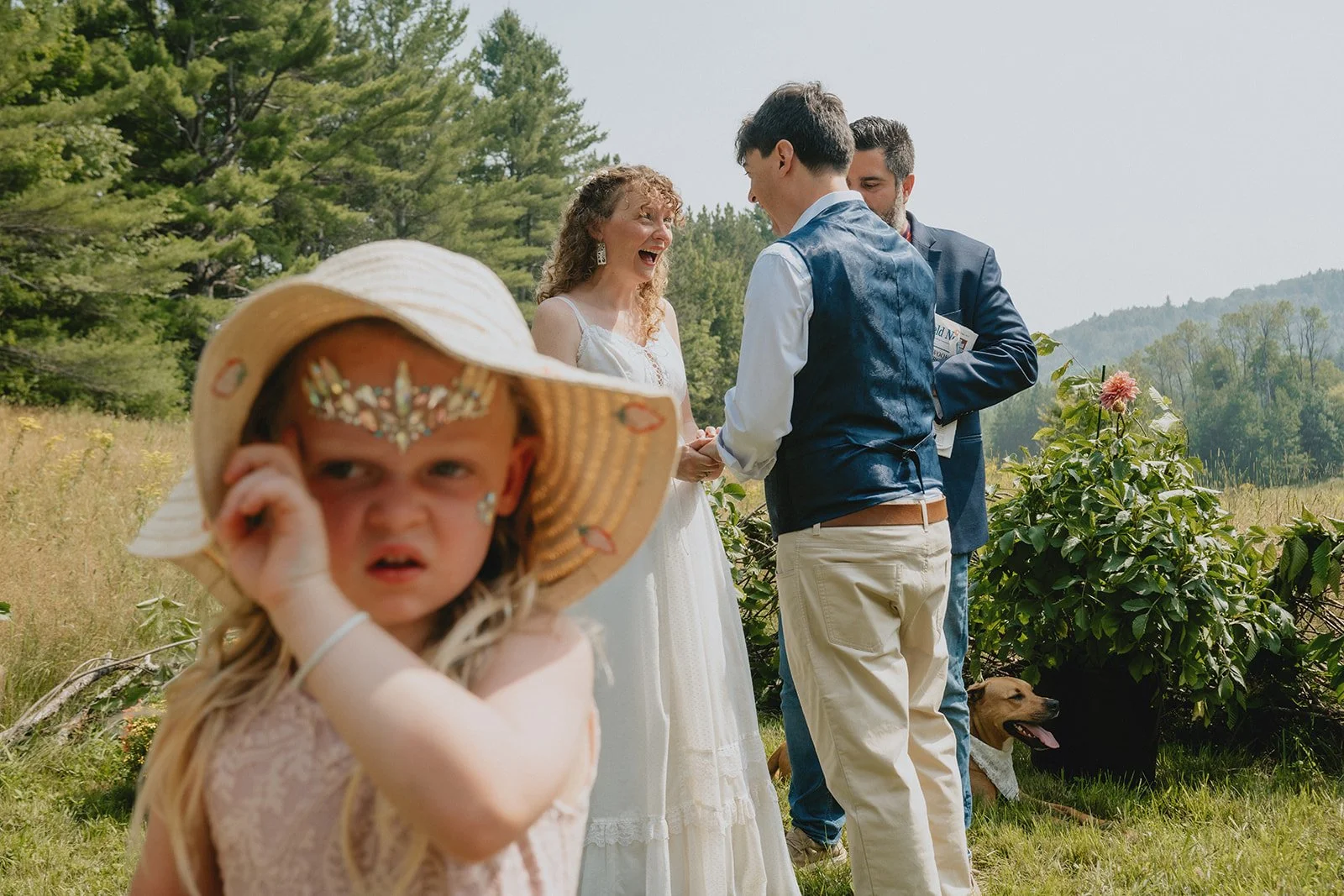 A young girl with a worried expression, wearing a hat and decorative face jewelry, in the foreground. In the background, a wedding ceremony taking place outdoors with a bride and groom exchanging vows, surrounded by scenic nature and a couple of dogs