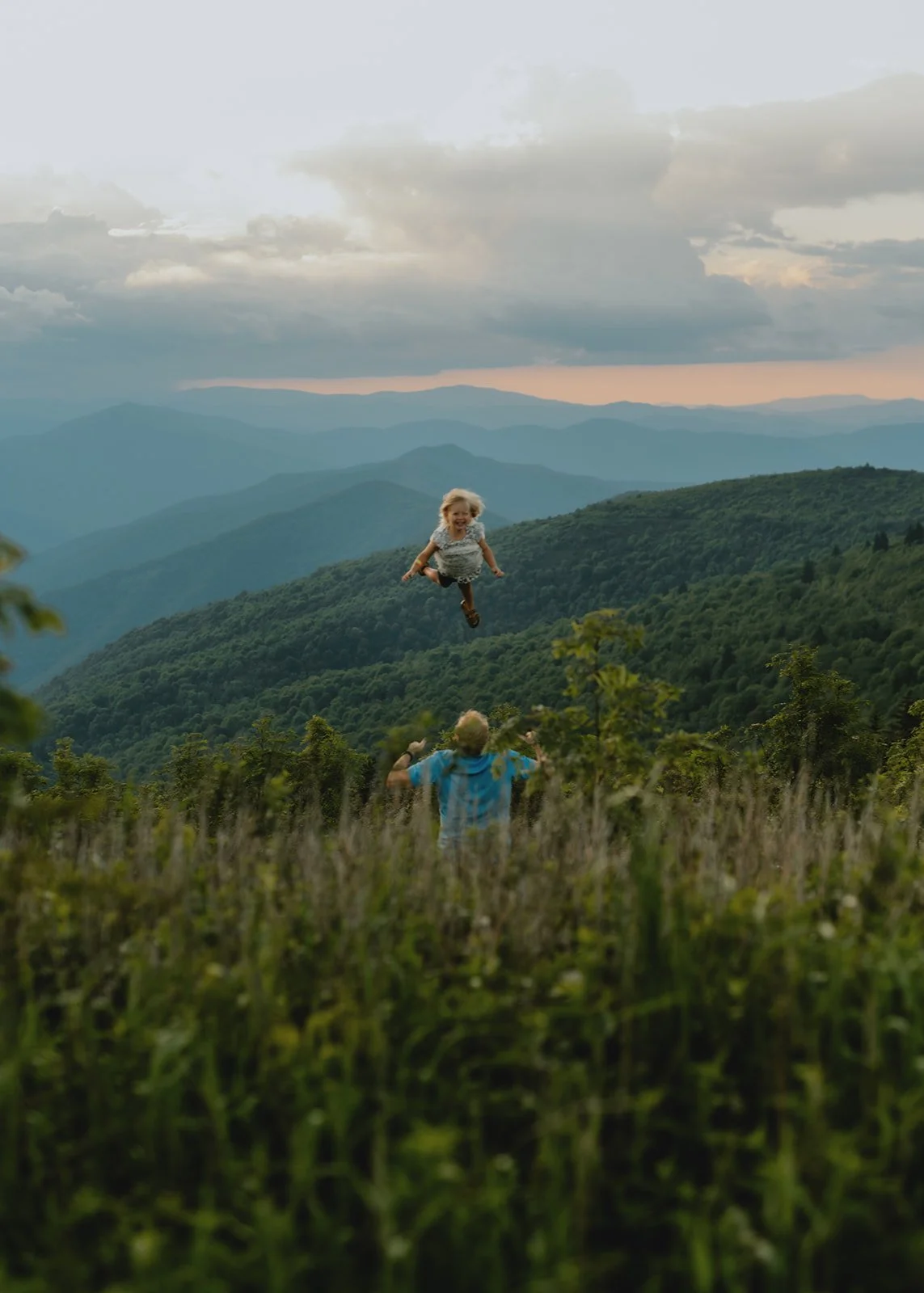 A young girl flying through the air, held by a man in a field of tall grass, with a backdrop of mountains and a cloudy sky at sunset. This is a picture taken my Photographer Melina Coogan on the mountaintop photography of Black Balsam Knob in NC.