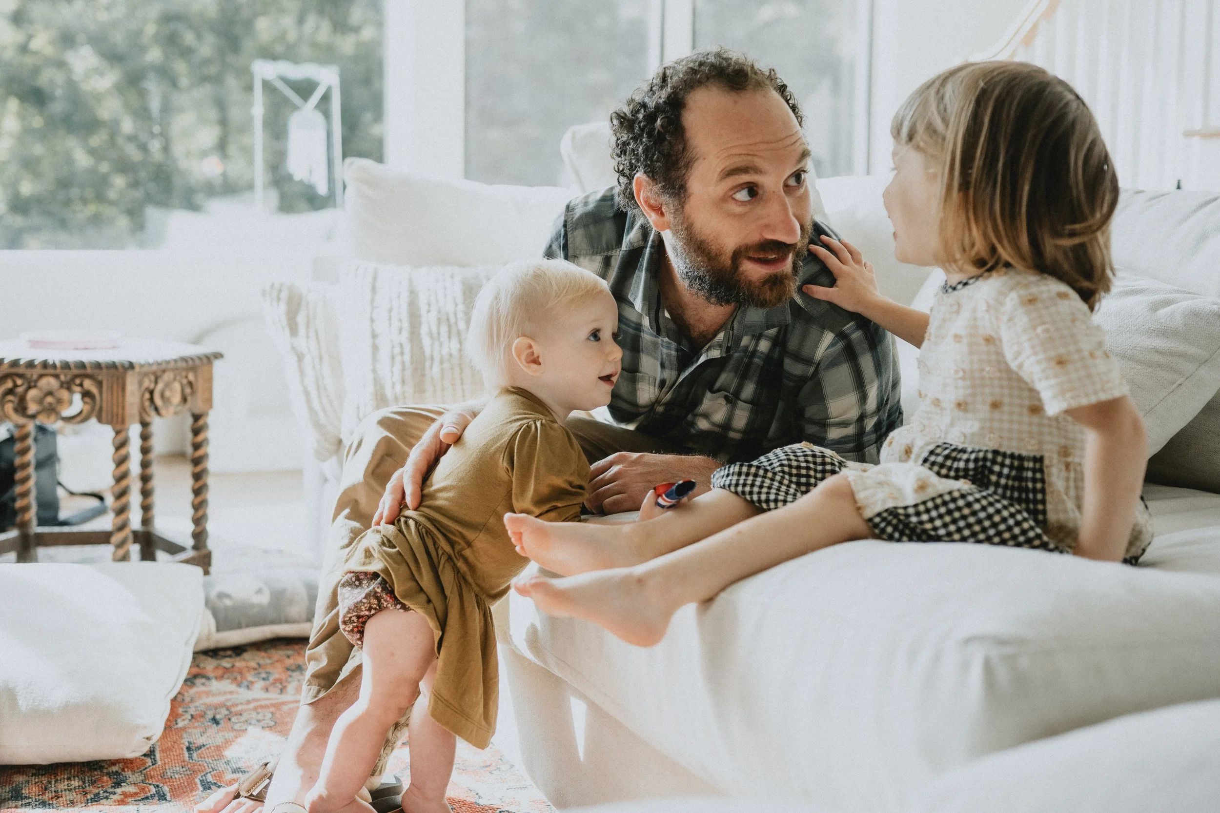 A man with curly hair and a beard is lying on a sofa, talking to two young children, a girl and a boy, in a bright, cozy living room with large windows and decorative furniture.
