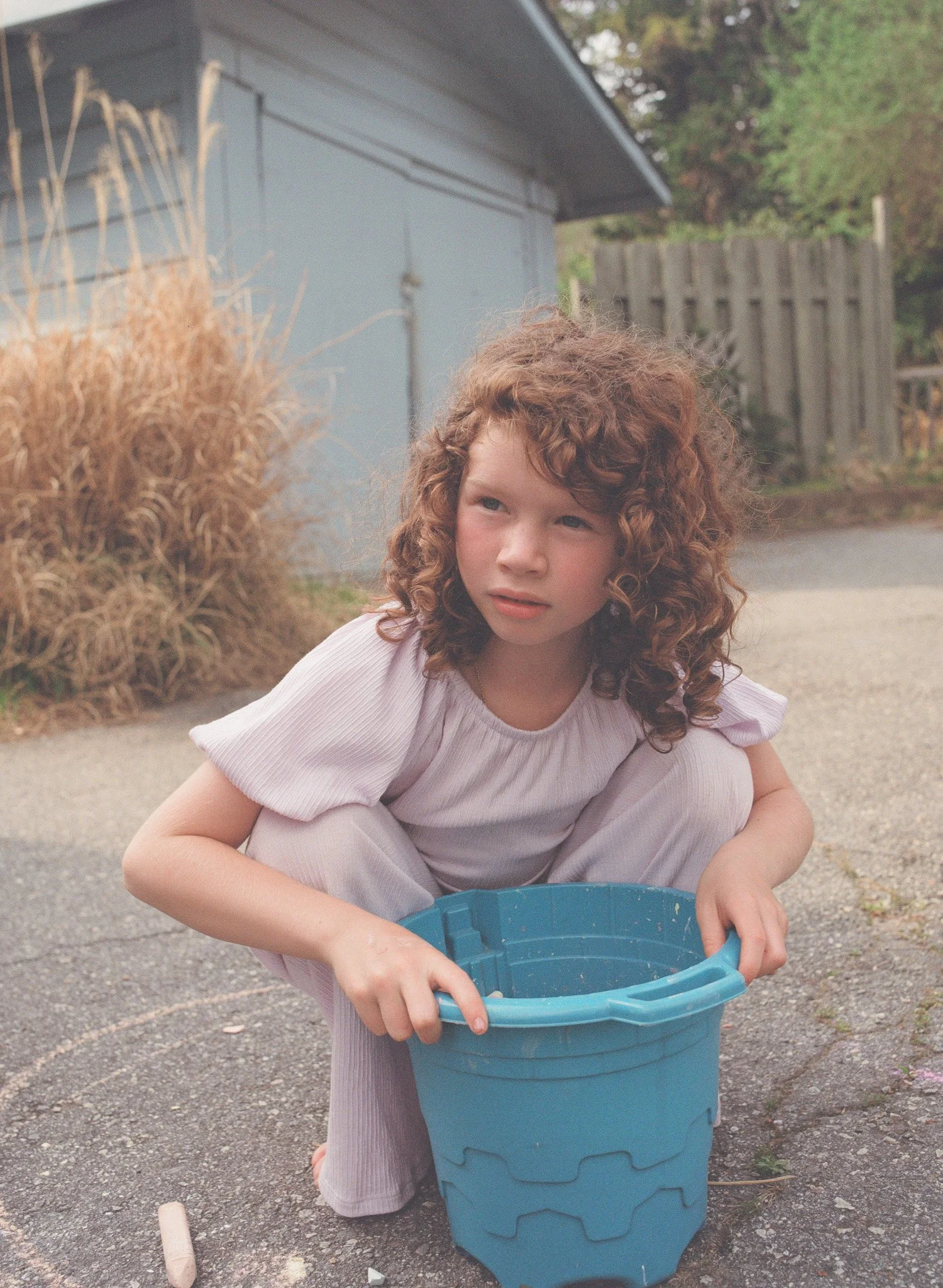 An eight year old girl with curly hair wearing a lavender colored jumpsuit pauses over a blue bucket of chalk. The picture is rendered in film and made in Asheville, North Carolina.