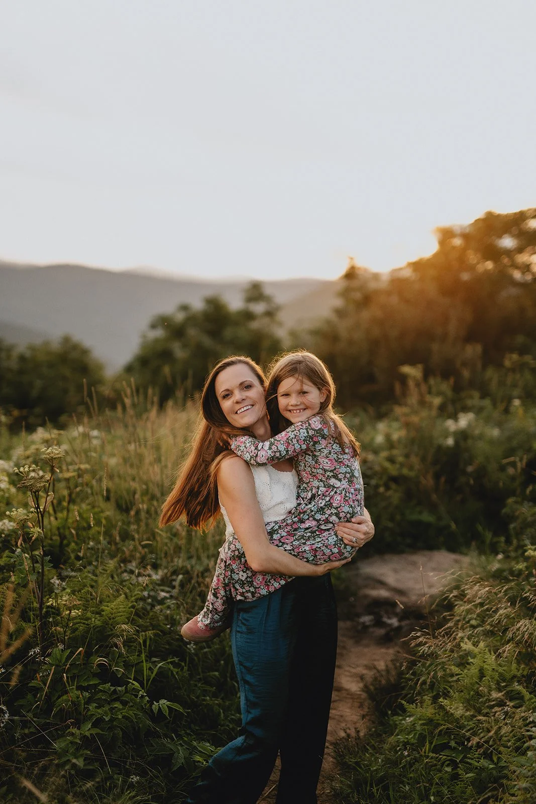 A woman holding a young girl outdoors during sunset, surrounded by tall grass and trees with mountains in the distance.