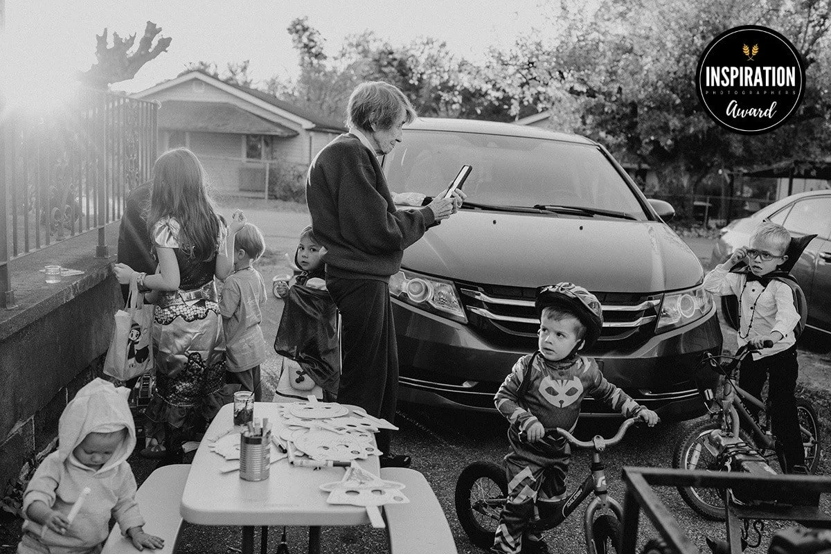 A black and white photo showing a woman and children gathered outdoors by a parked car, with some children wearing helmets and holding bikes, and a table with various items. The woman is looking at a tablet. There is a badge reading 'Inspiration Awar
