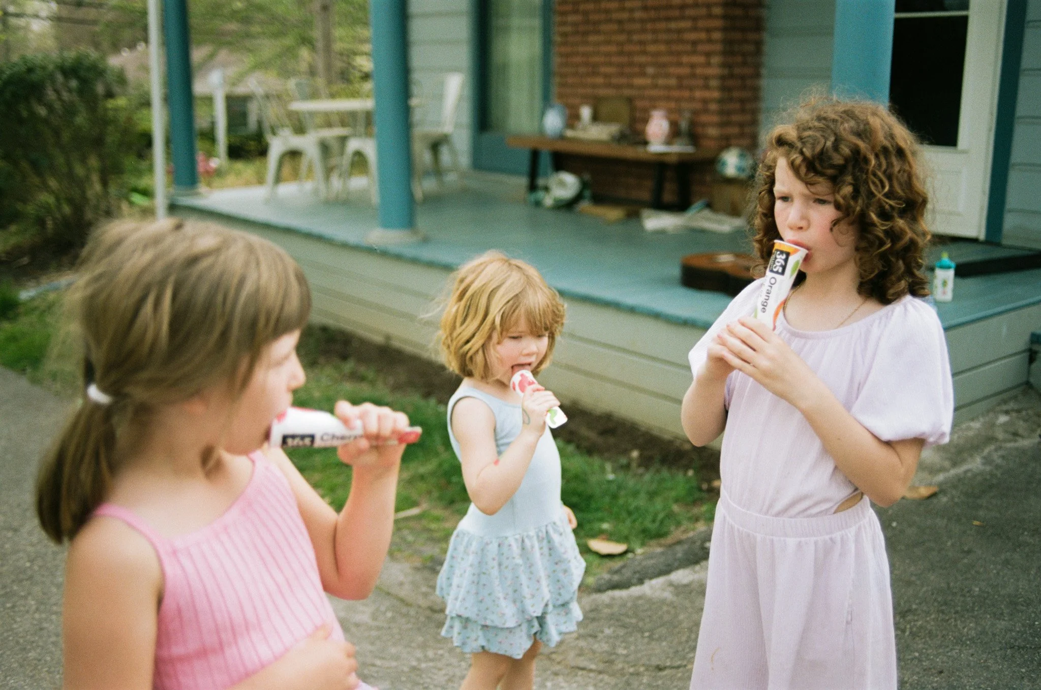 Three young girls drink popsicles in their backyard. They are wearing pastel, easter colored dresses. The mood is quiet, mellow and intimate.