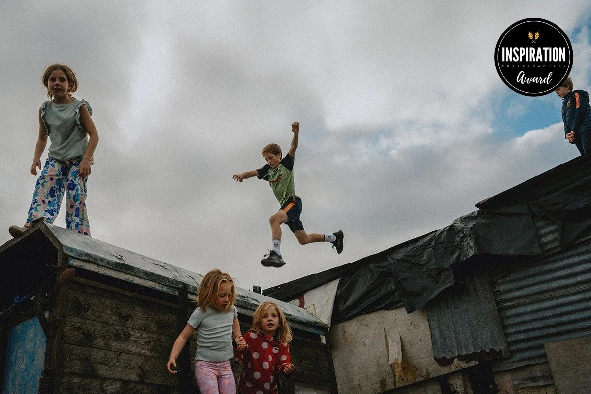 Children playing and jumping on a makeshift playground made of wooden and metal structures outdoors on a cloudy day.