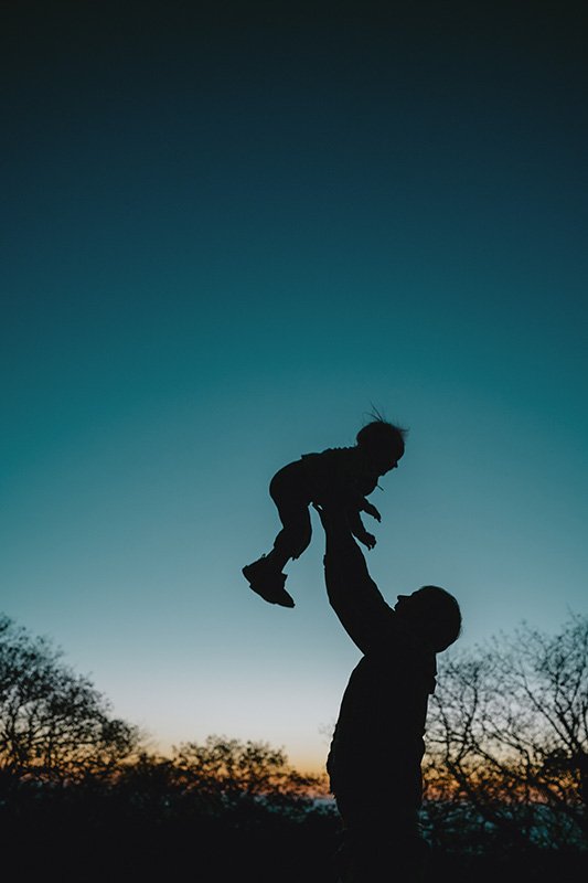 A silhouetted child is tossed into the air by her dad against a blue sky in the mountains of asheville during a family photo shoot