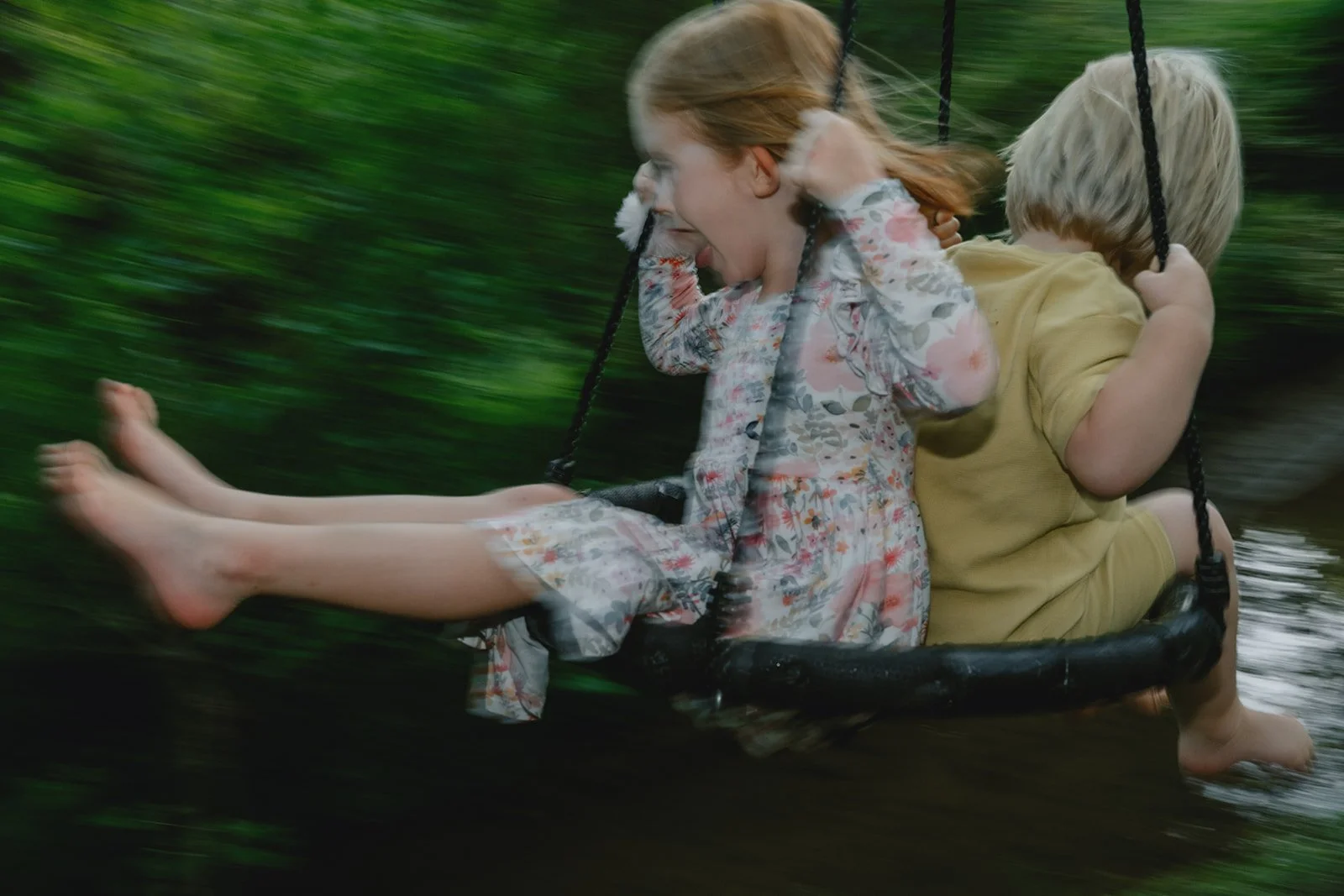 a brother and sister with red and blonde hair swing on a swing with a blurry green background that demonstrates the movement.