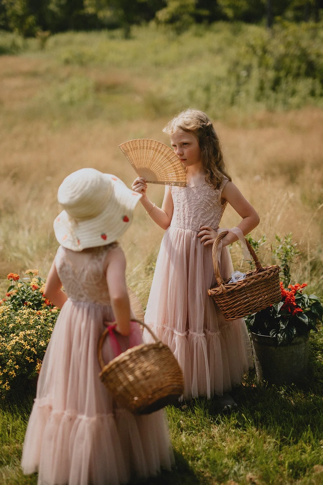 Two young girls in pink dresses and sun hats carrying baskets and facing each other in a grassy outdoor setting with flowers.