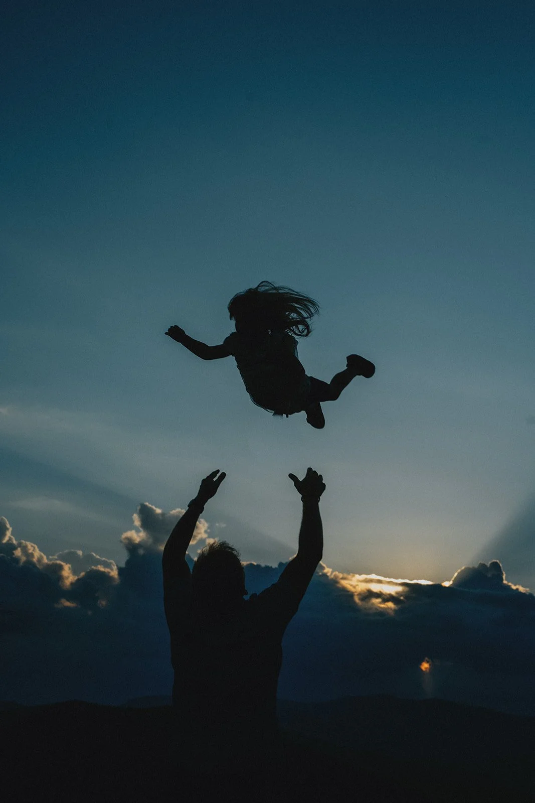 Silhouette of a man tossing a girl in the air outdoors during sunset or dusk, with clouds and mountains in the background.