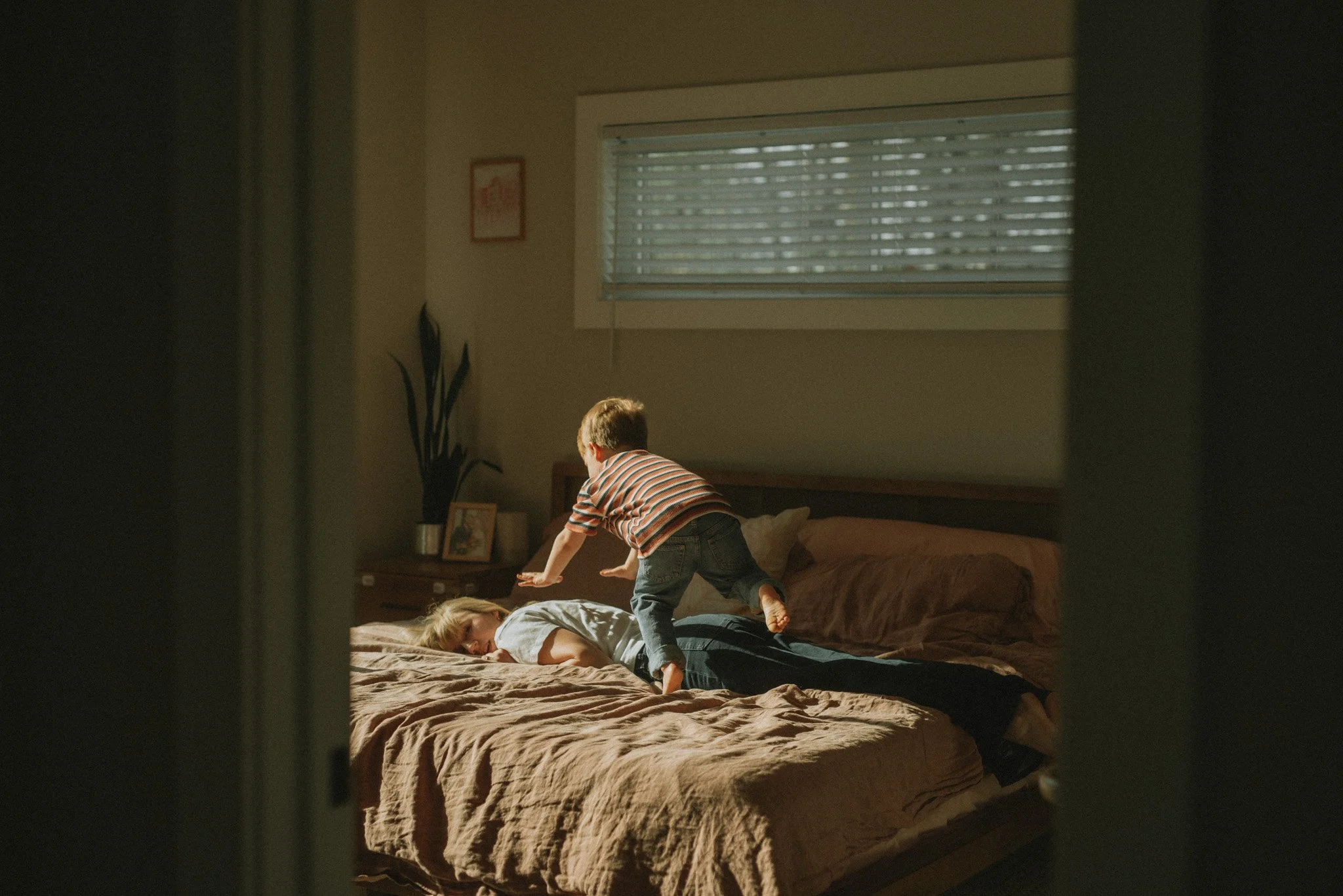 The photo is looking in on a bedroom scene in low-light. An exhausted mother lies belly first on the bed with her head turned to the side. A three year old child is jumping onto her, and the photographer captured the child mid-jump in the air.