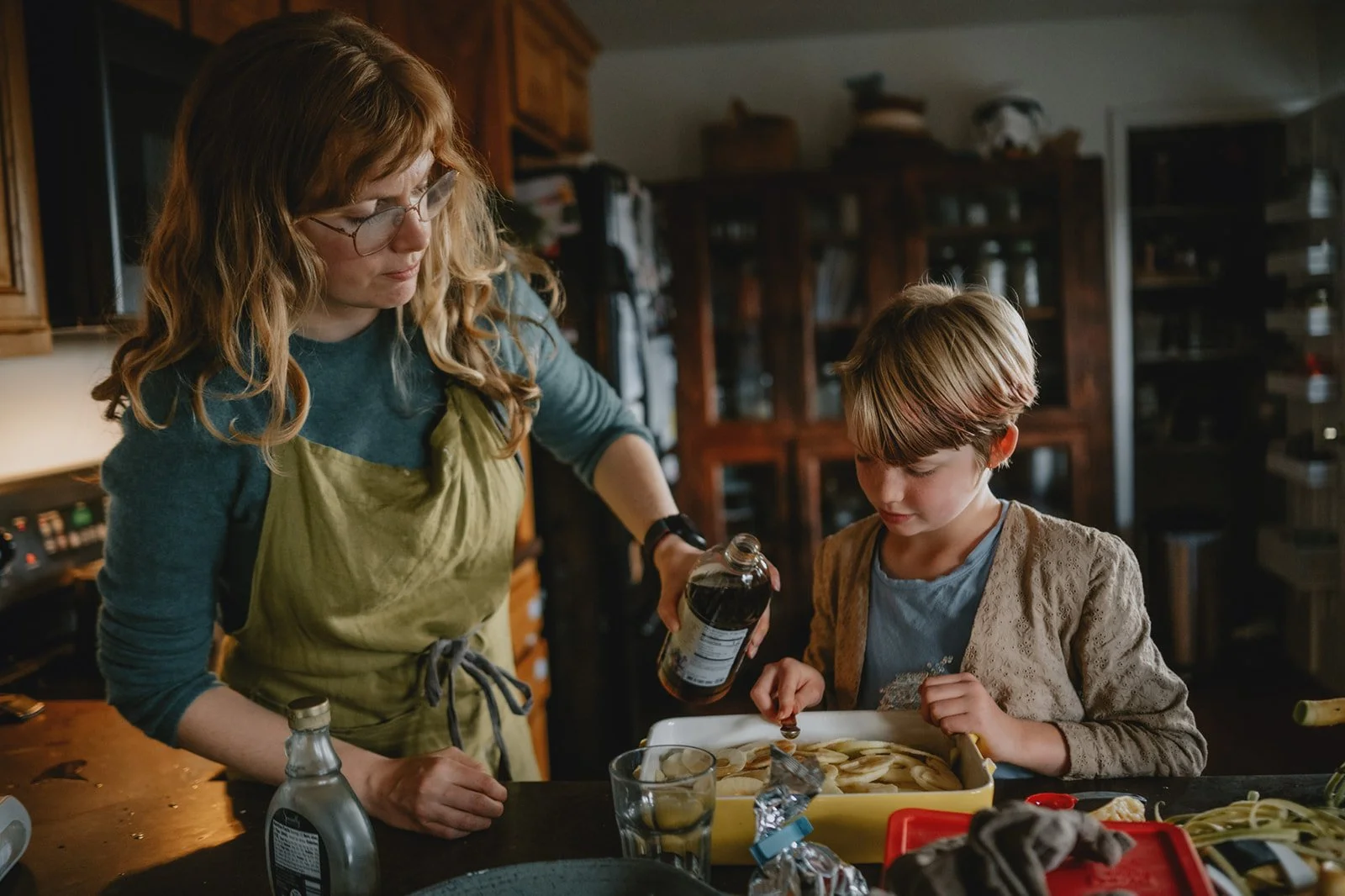 A woman with glasses and wavy red hair, wearing a blue shirt and green apron, is pouring liquid from a bottle over a tray of sliced bananas, while a young blonde girl with a short haircut and a beige jacket watches attentively in a kitchen.