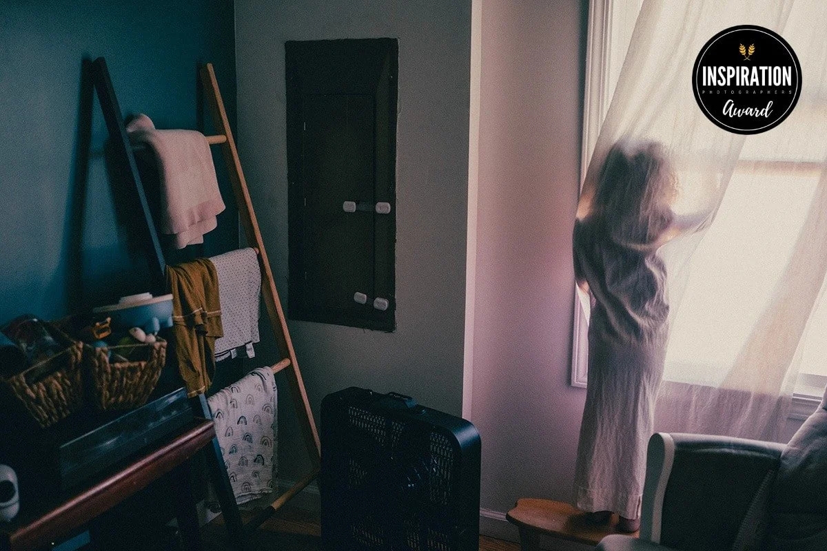 A woman standing by a window with sheer curtains, pulling them open to let in light.