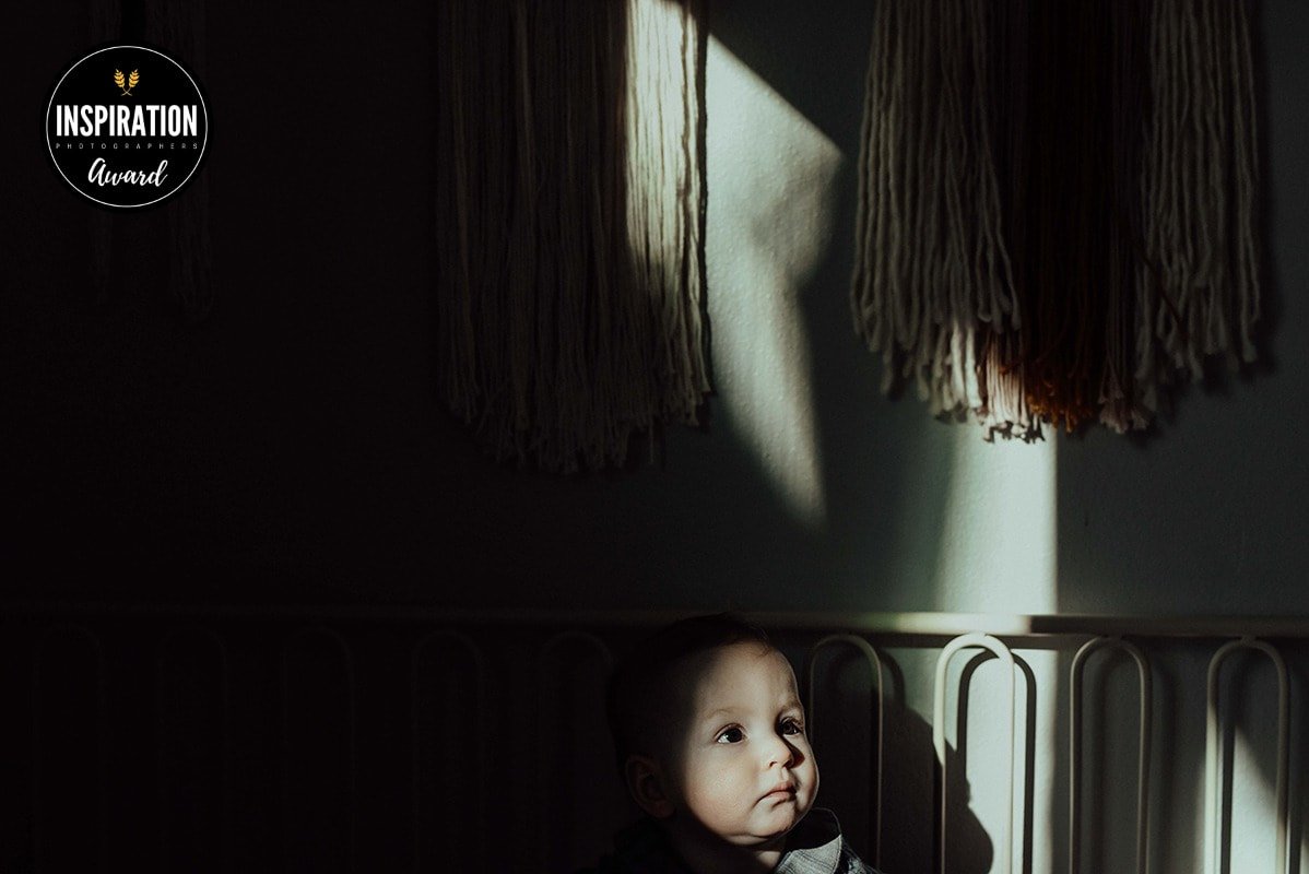 A toddler sitting on a bed in a dark room, with sunlight casting a star-shaped shadow on the wall behind him.