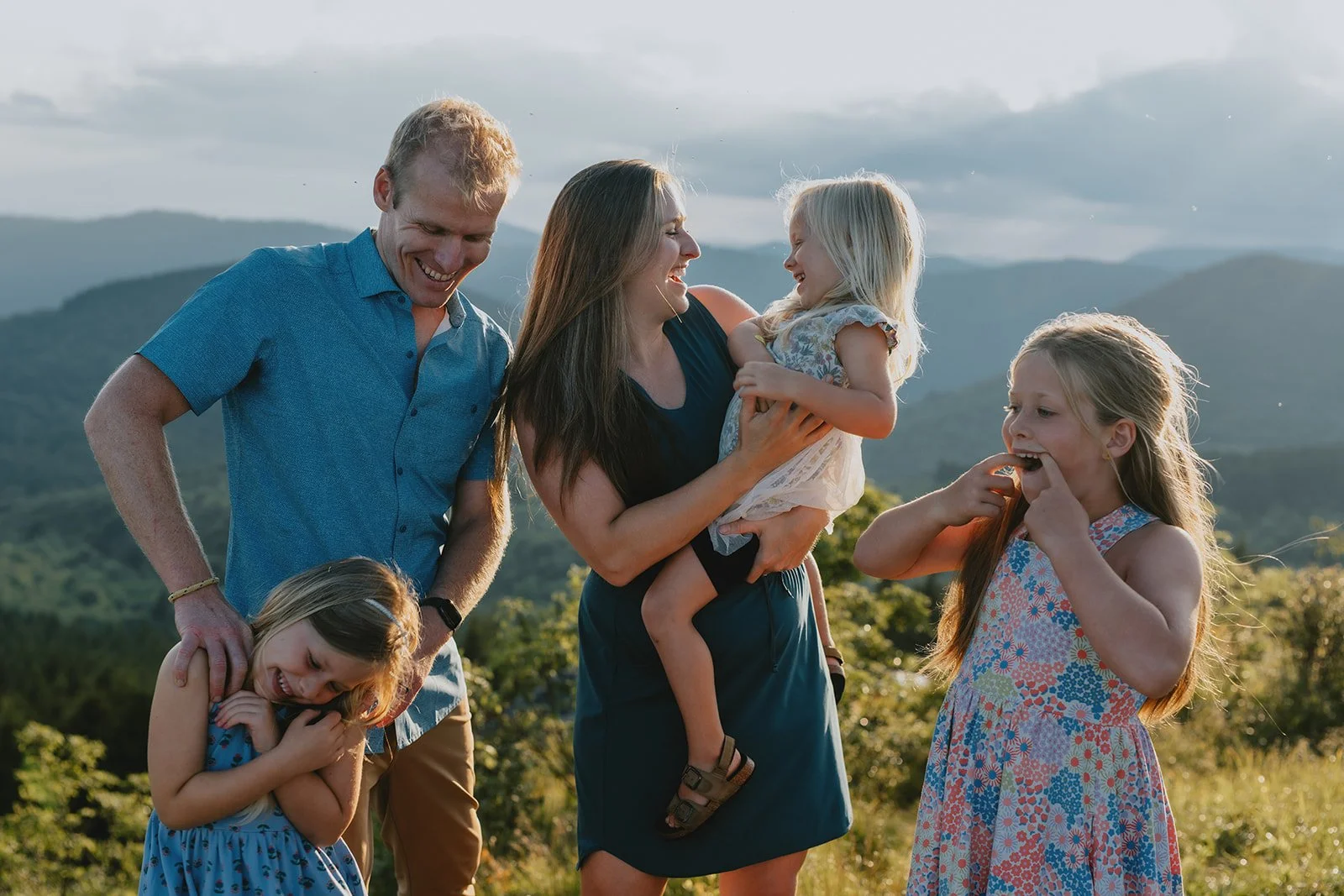 A family of five, including two adult women, one adult man, and three young girls, enjoying a moment outdoors in a scenic landscape with mountains. They are smiling, laughing, and engaging with each other, with one girl being held and the others play