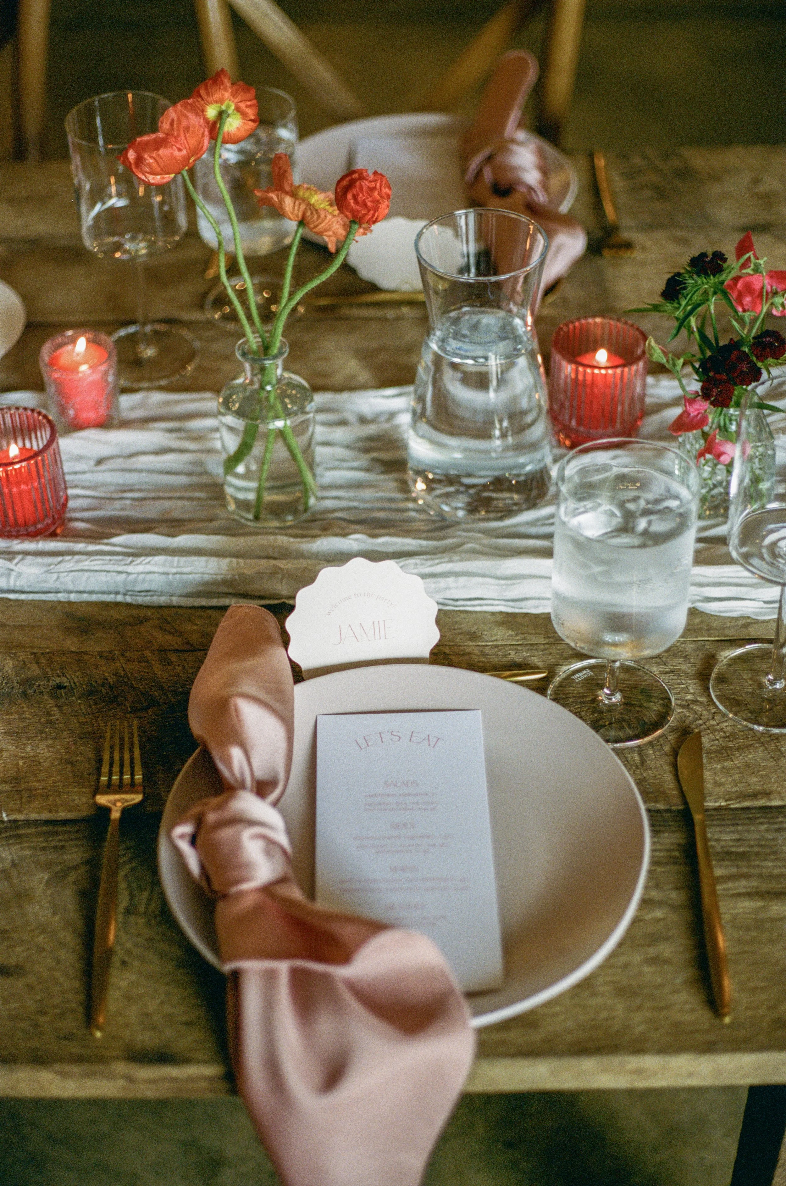 A decorated table set for a special event with flowers, candles, glassware, and a menu, featuring a place card with the name 'Jamie'.