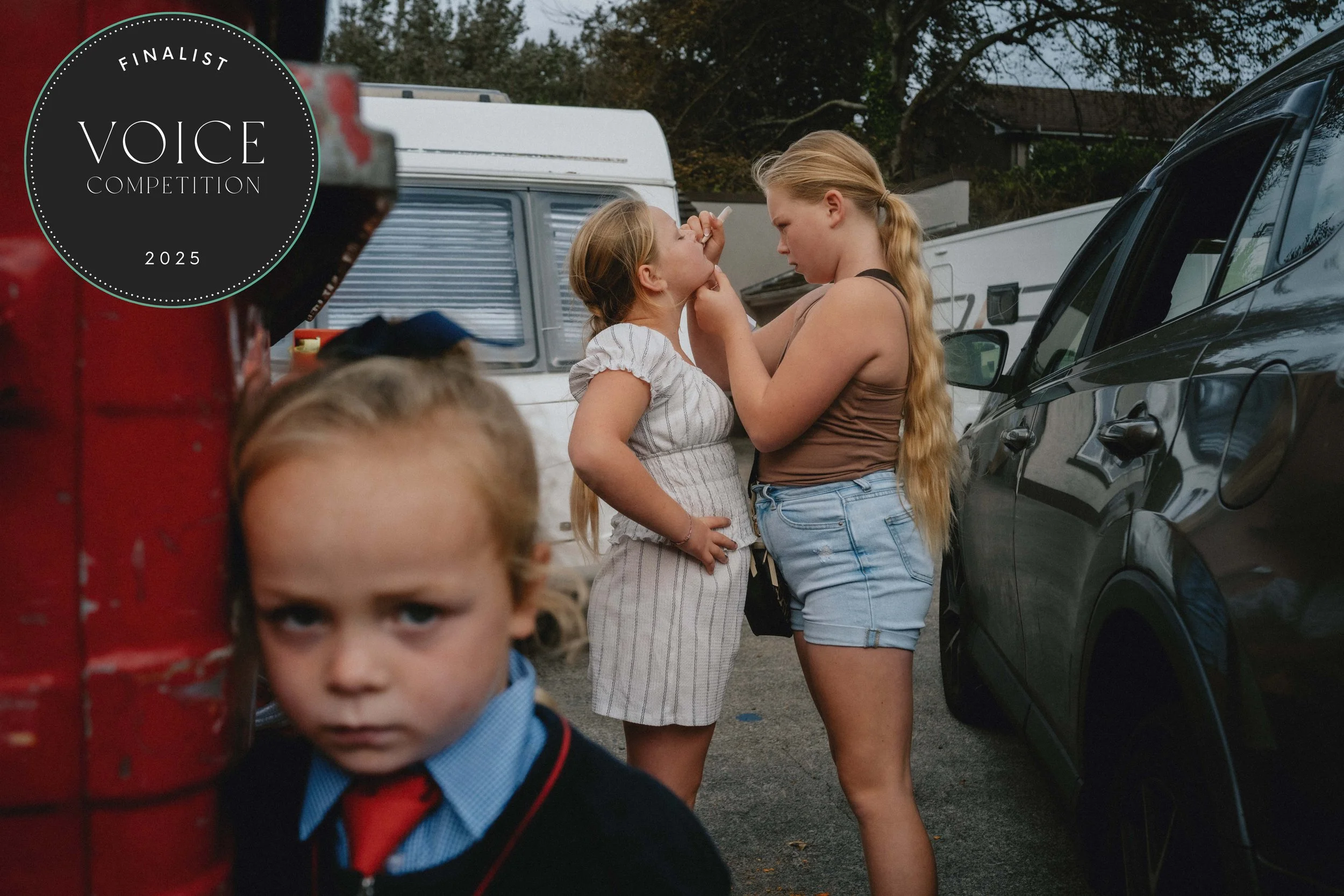 A woman and a young girl engaging in a close, intimate moment outdoors near cars, with a young boy in the foreground looking at the camera, and a black sticker on the top left corner reading 'Finalist Voice Competition 2025'.