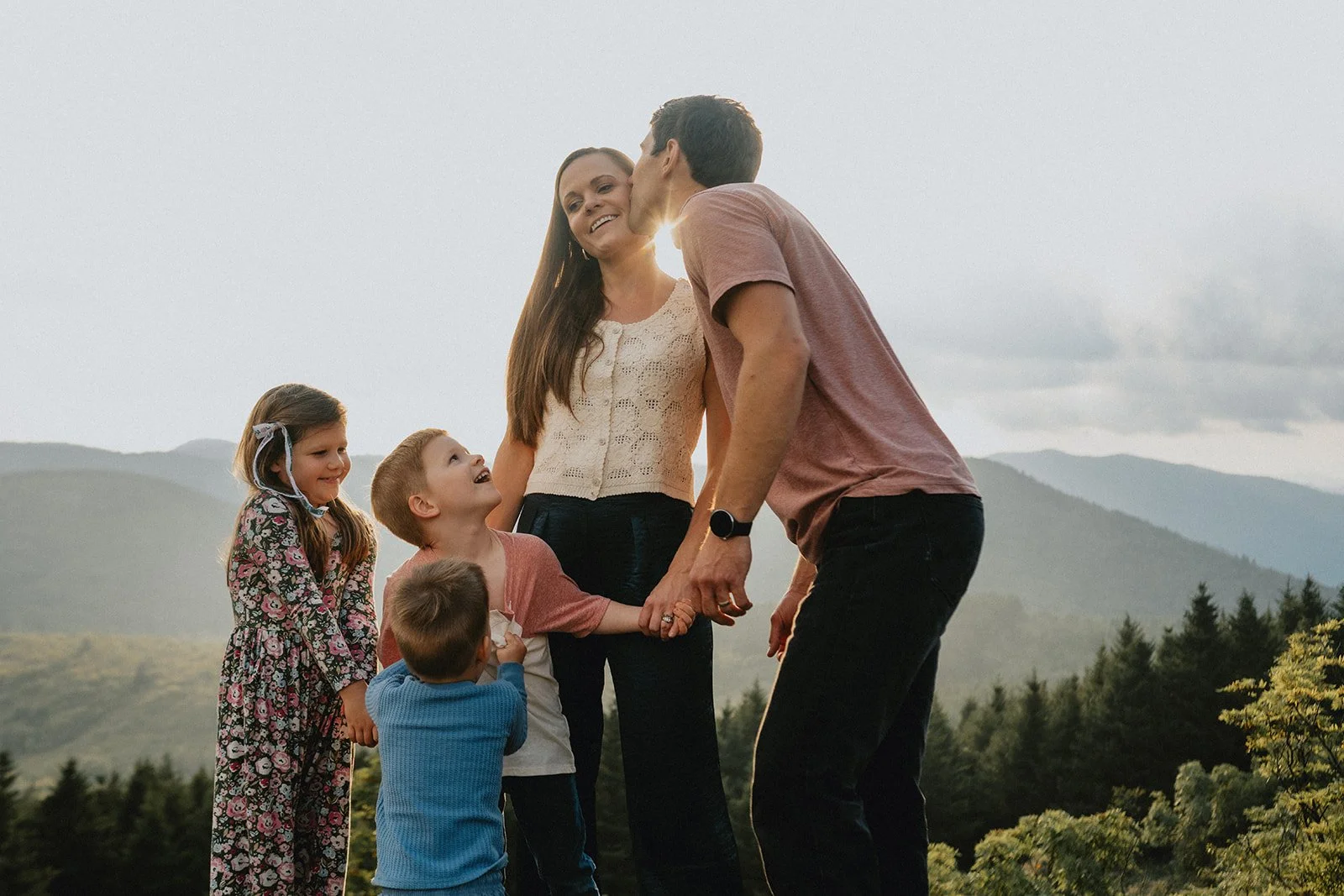 A family of five holding hands outdoors in front of mountains and trees, smiling and enjoying a moment together.