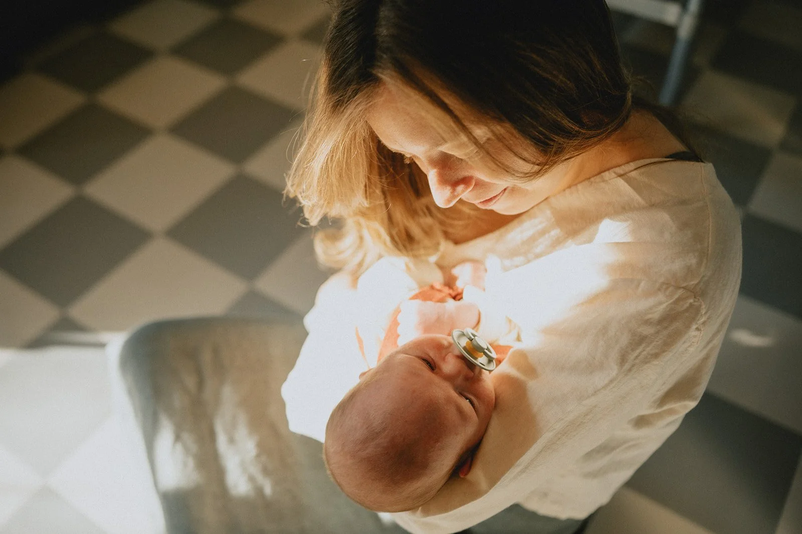 A woman holding a newborn baby, who has a pacifier in their mouth, in her arms. They are indoors with a checkered black and white floor.
