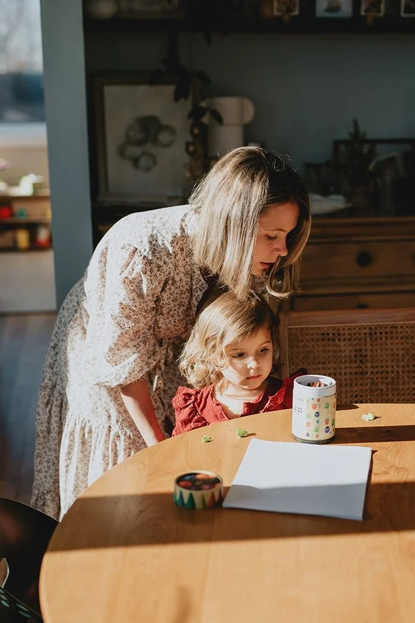 A mom helps her toddler girl get settled at a table to do some coloring while mom cooks, during an unposed family photography session in North Carolina