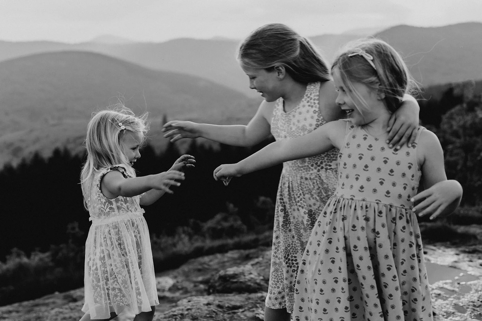 Three young girls playing joyfully on a rocky landscape in front of mountains, wearing dresses, with the eldest hugging the middle girl, and the youngest reaching out to the smallest, all smiling in black and white.