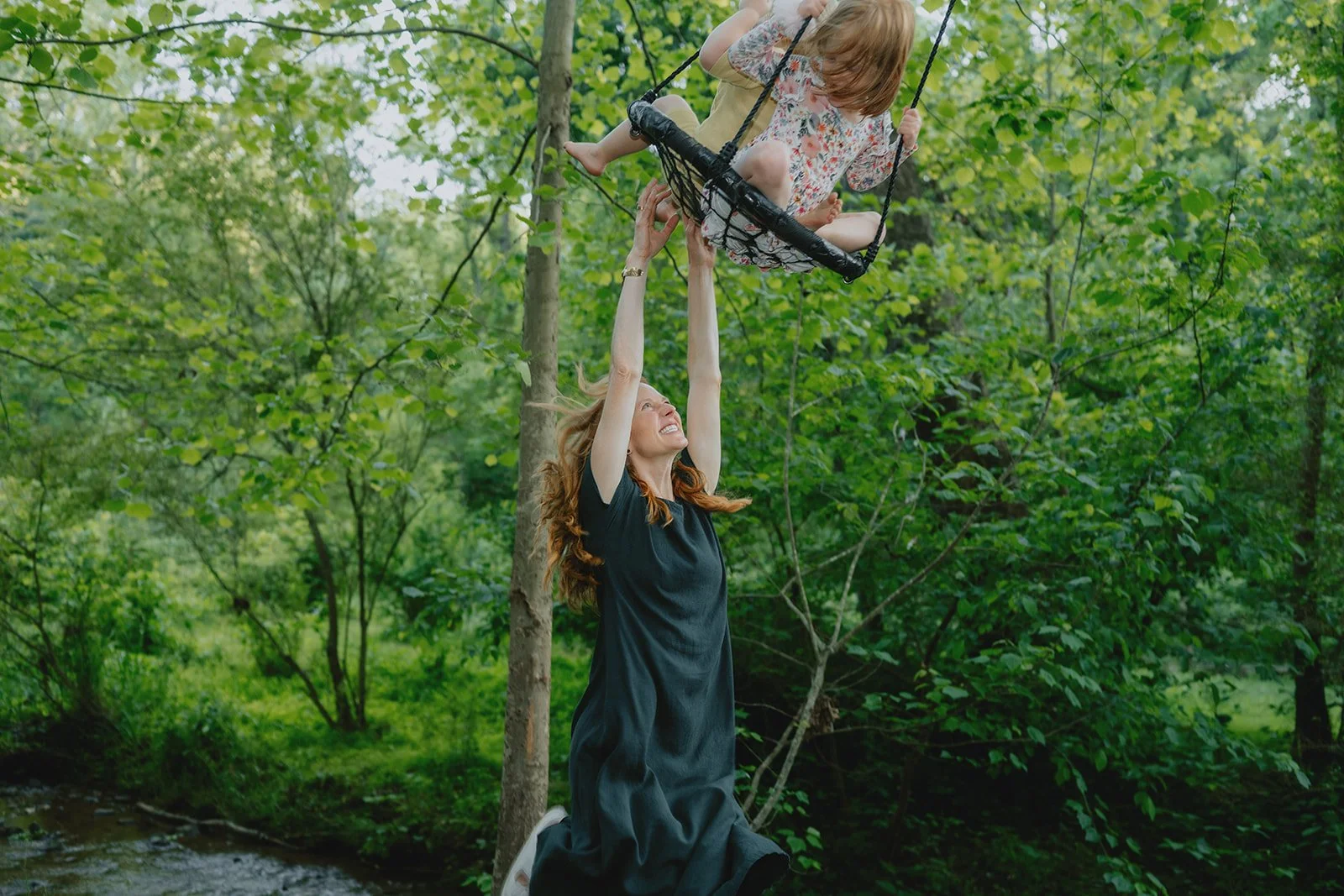 Woman kneeling on grass beneath a tree, holding a young girl on a swing in a lush green outdoor setting.