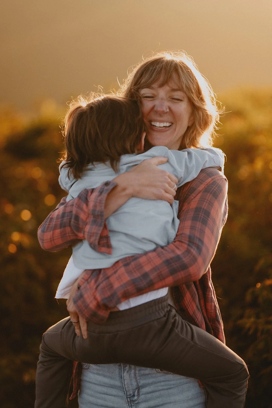 A woman with curly hair is happily hugging a young boy, who is sitting on her hip. They are outdoors during sunset, with warm lighting and blurred trees in the background. It was made by the best family photographer in Asheville, NC.