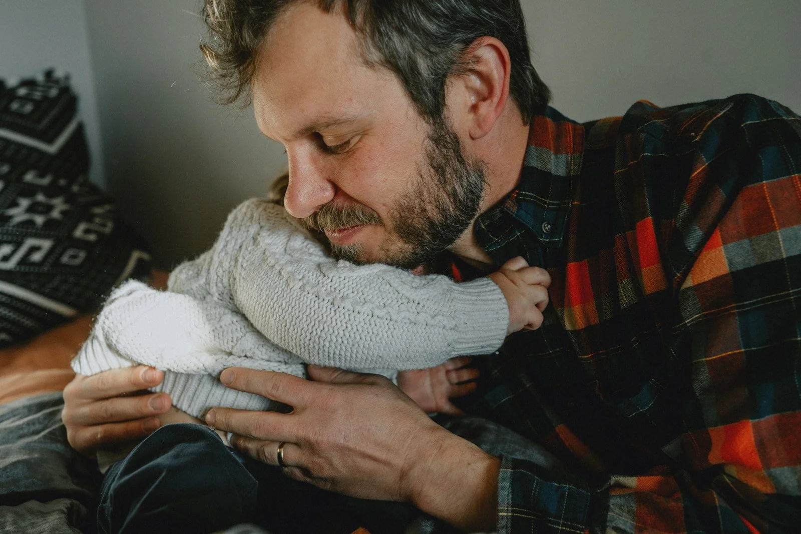 A man with a beard and a plaid shirt holds a young child, possibly a toddler, close to his face. The child is wearing a white knitted sweater and is nestled against the man's chest, creating an intimate and tender moment.