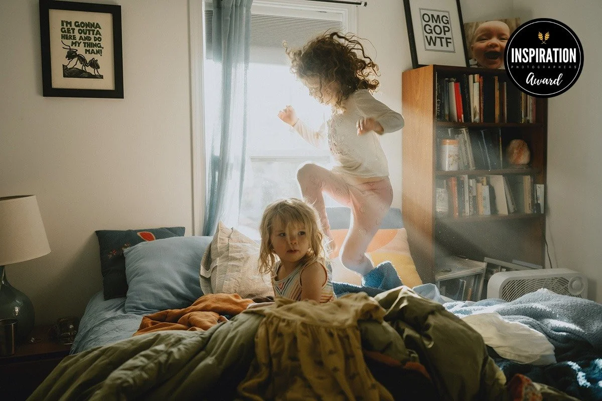 Two young girls jumping on a bed in a bedroom illuminated by natural light. One girl with blonde hair looks surprised while the other with curly hair is mid-air.