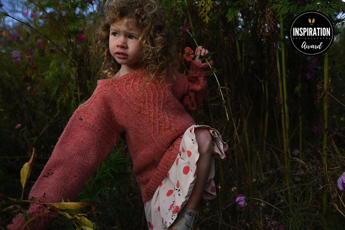 A young girl with curly hair and fair skin wearing an oversized pink sweater and a white skirt with red and pink polka dots, standing in a dense, dark green outdoor area with purple flowers. She is holding a small flower in her right hand and looking