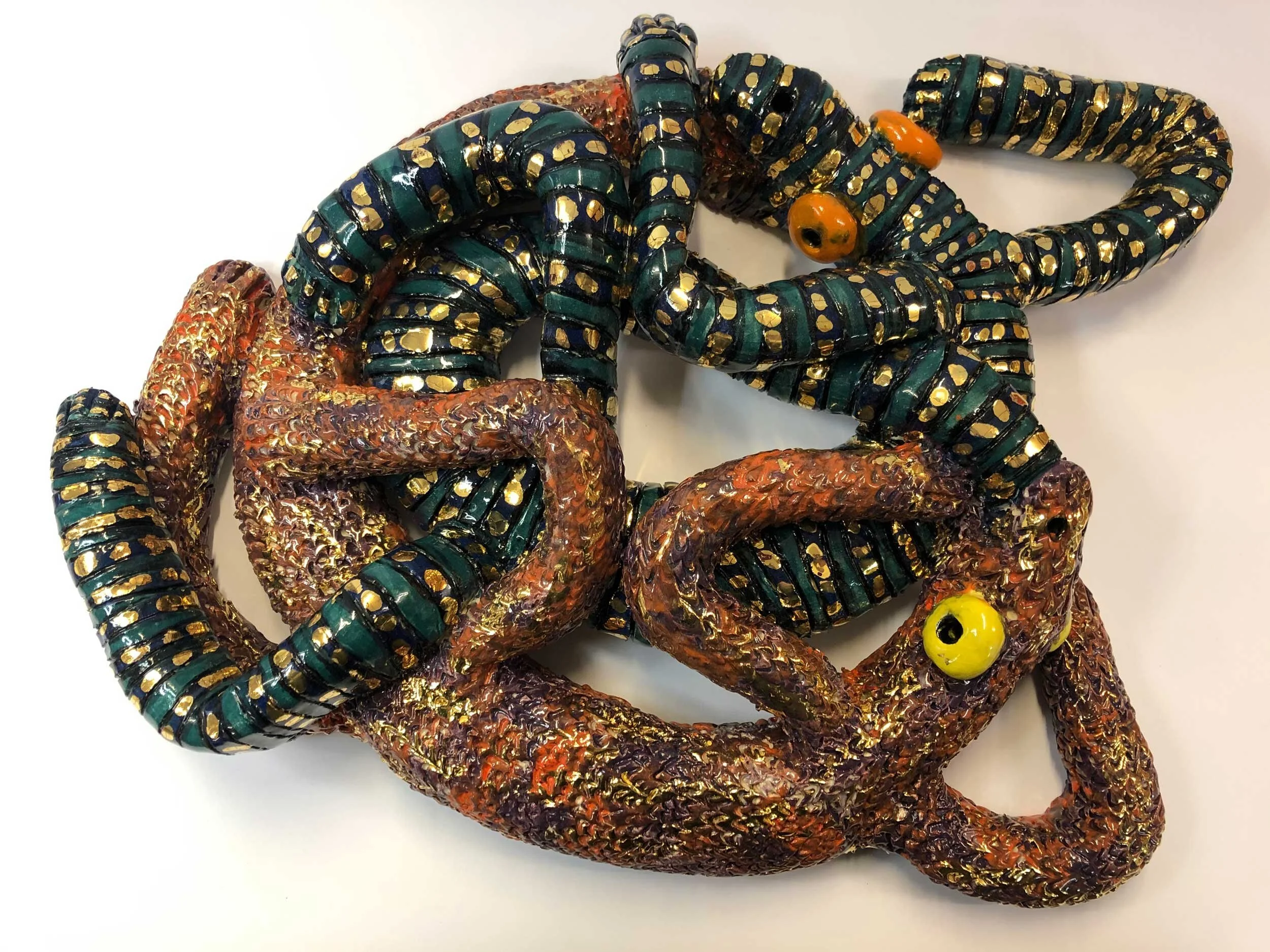 A ceramic piece with a golden shine over the top. Two lizards (one brown/orange and the other green/black) are entwined together in a circular 3D form in clay. 